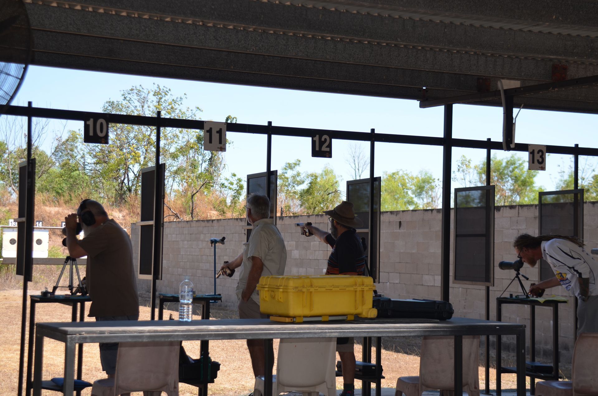 People shooting at a gun range. Targets, numbers 10-13. Sunny, outdoor setting.