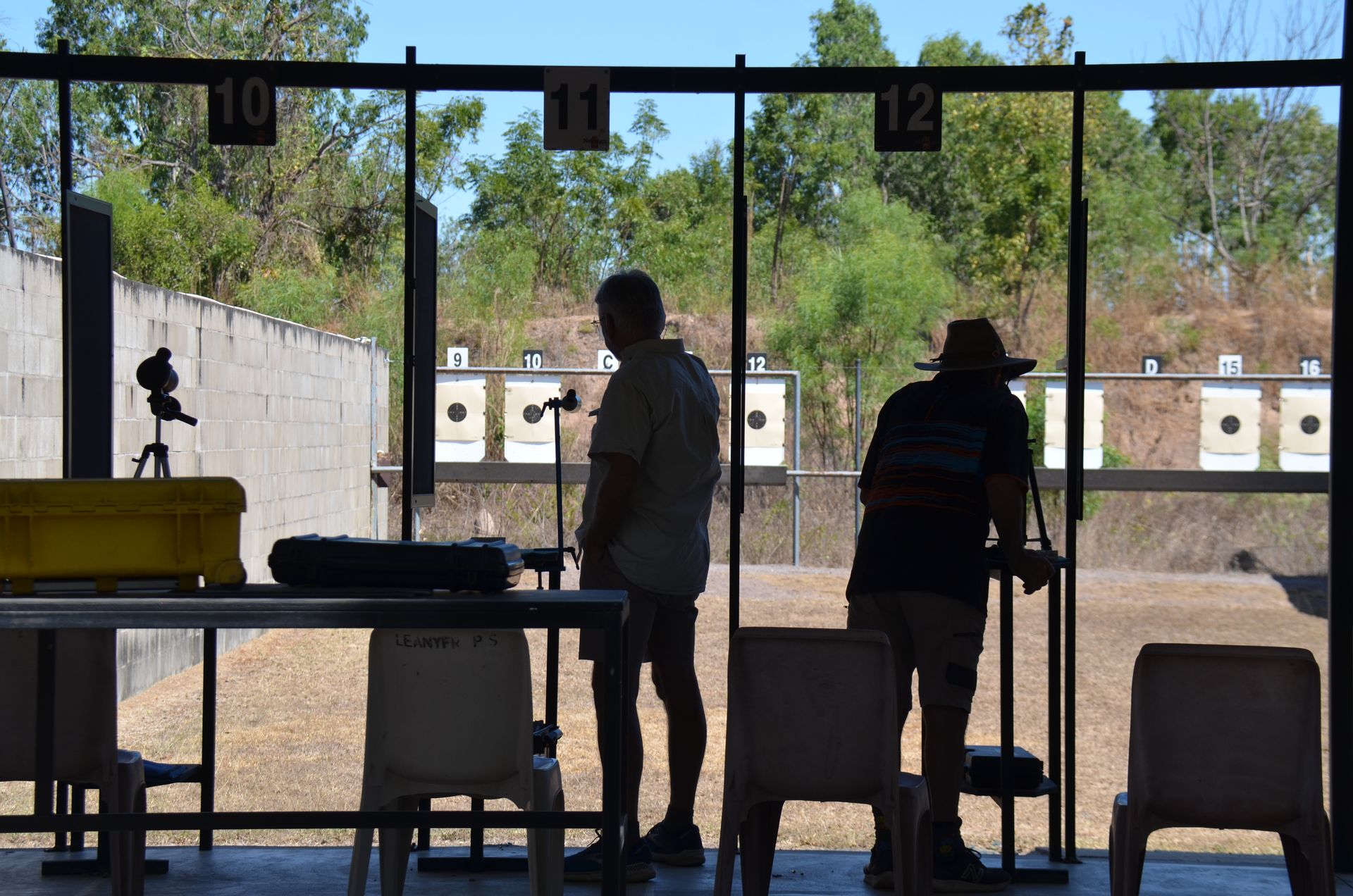 Two people at an outdoor shooting range, silhouetted. Targets in the background.