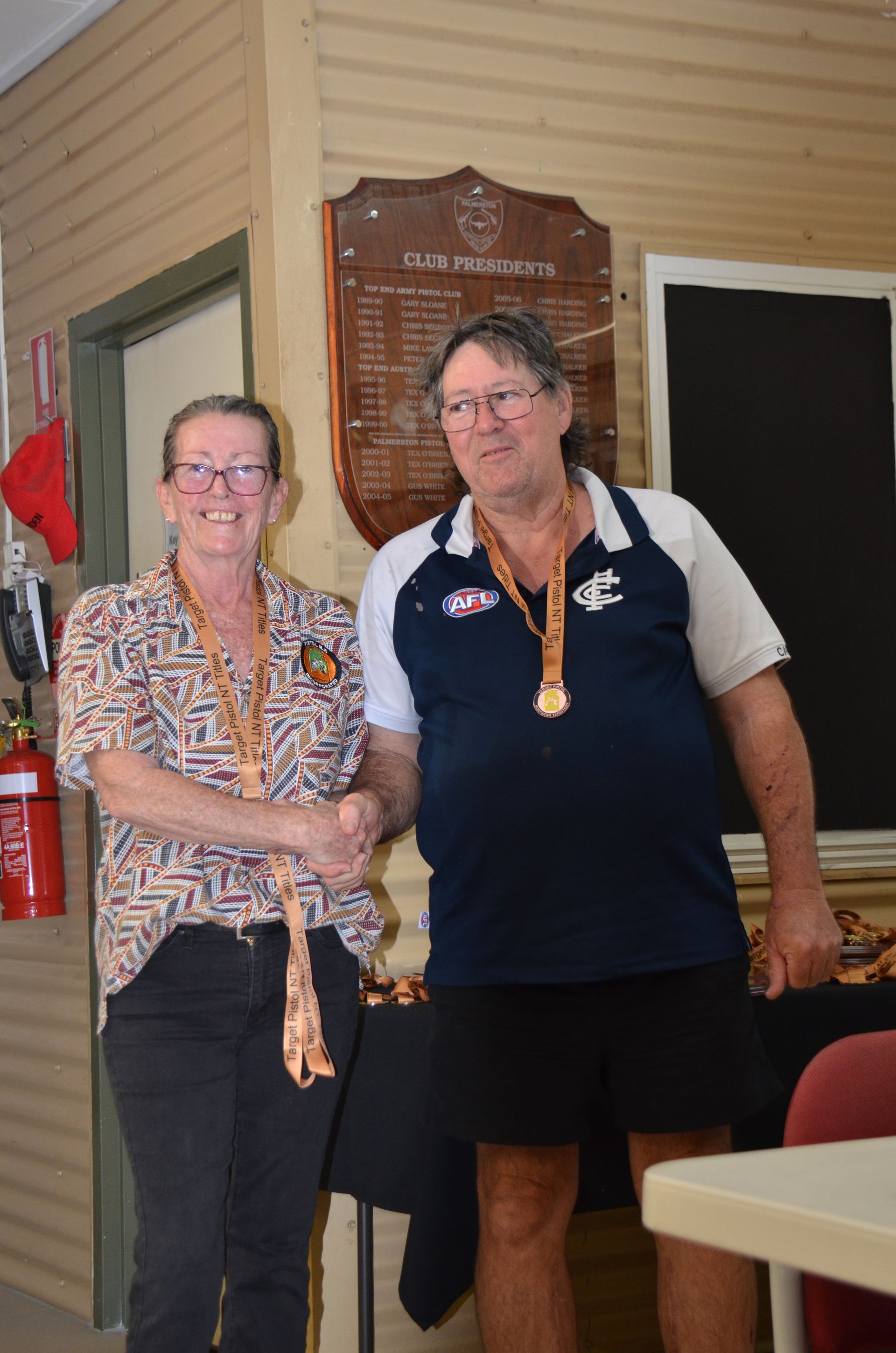 Two people with medals; woman in patterned shirt, man in navy shirt, next to trophy board.