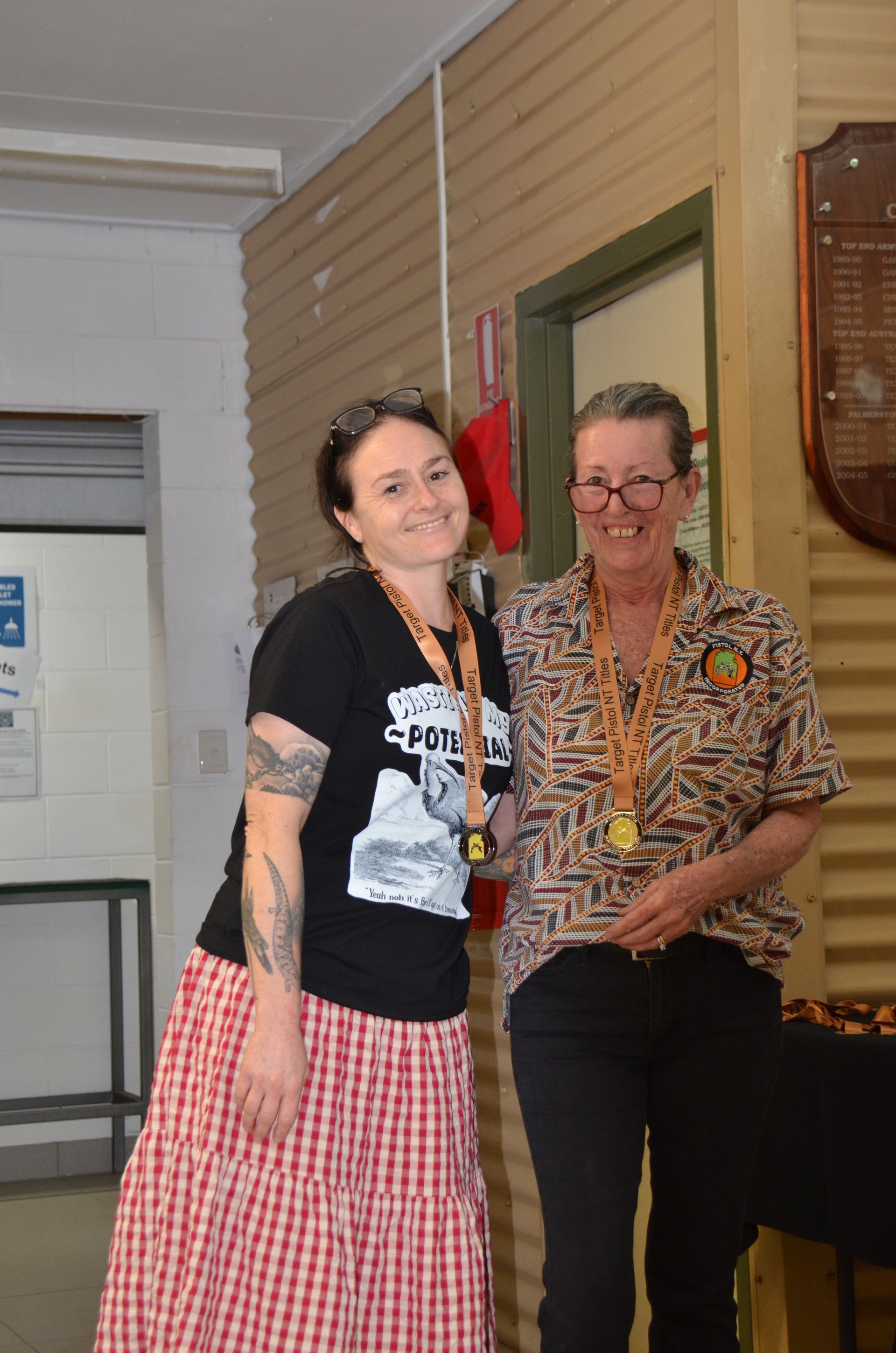 Two women smiling, wearing medals, in a building. One in black shirt, red skirt. The other in patterned shirt, black pants.