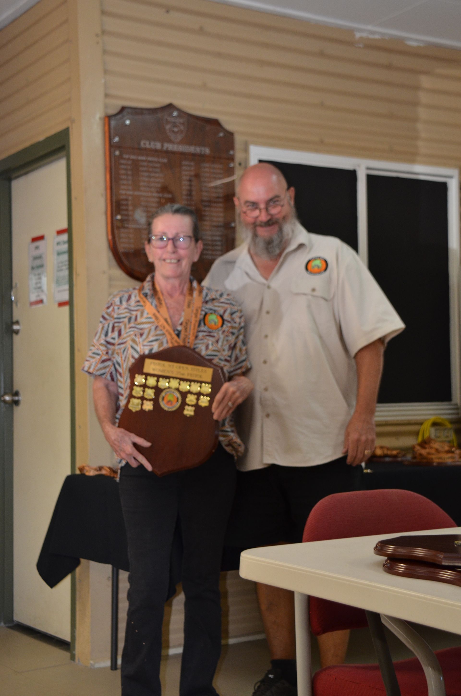 Woman and man smiling, holding a wooden trophy, in a room with awards on the wall.