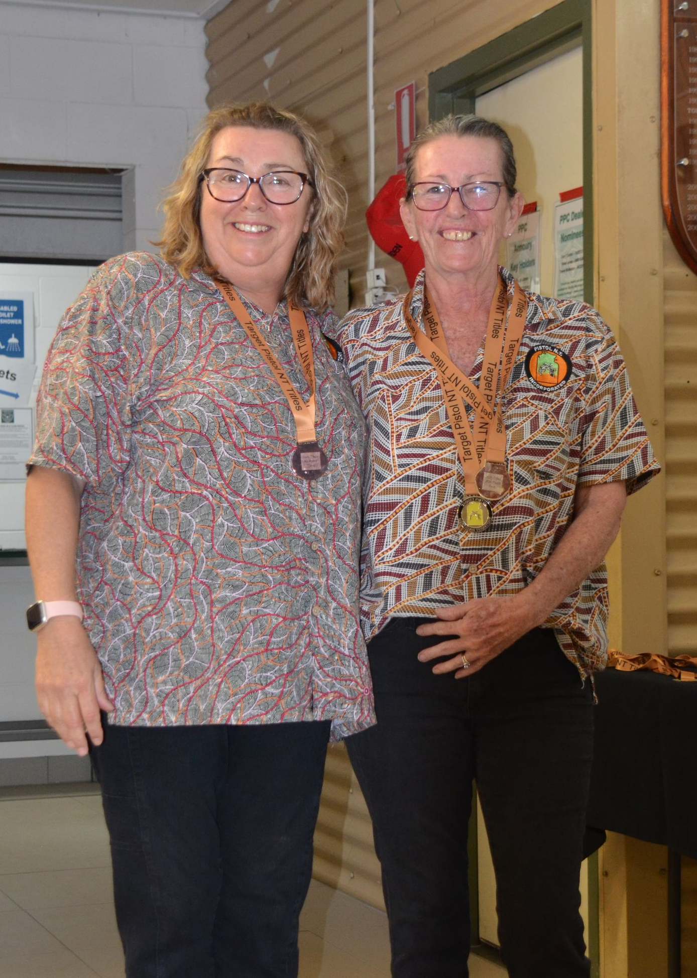 Two women smiling, wearing patterned shirts and medals, standing indoors.