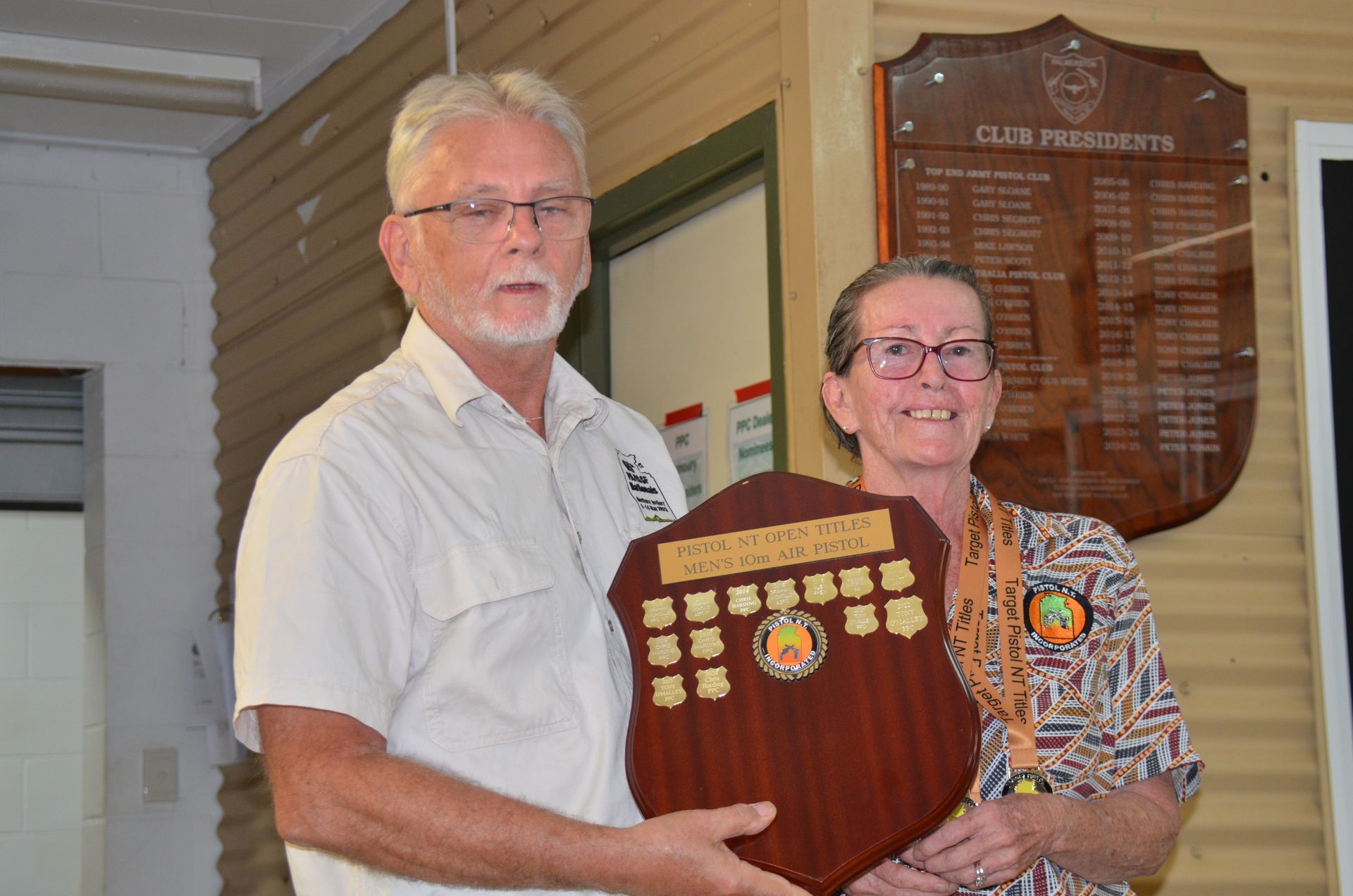 Man presenting a shield award to a woman indoors. Club Presidents plaque in background.