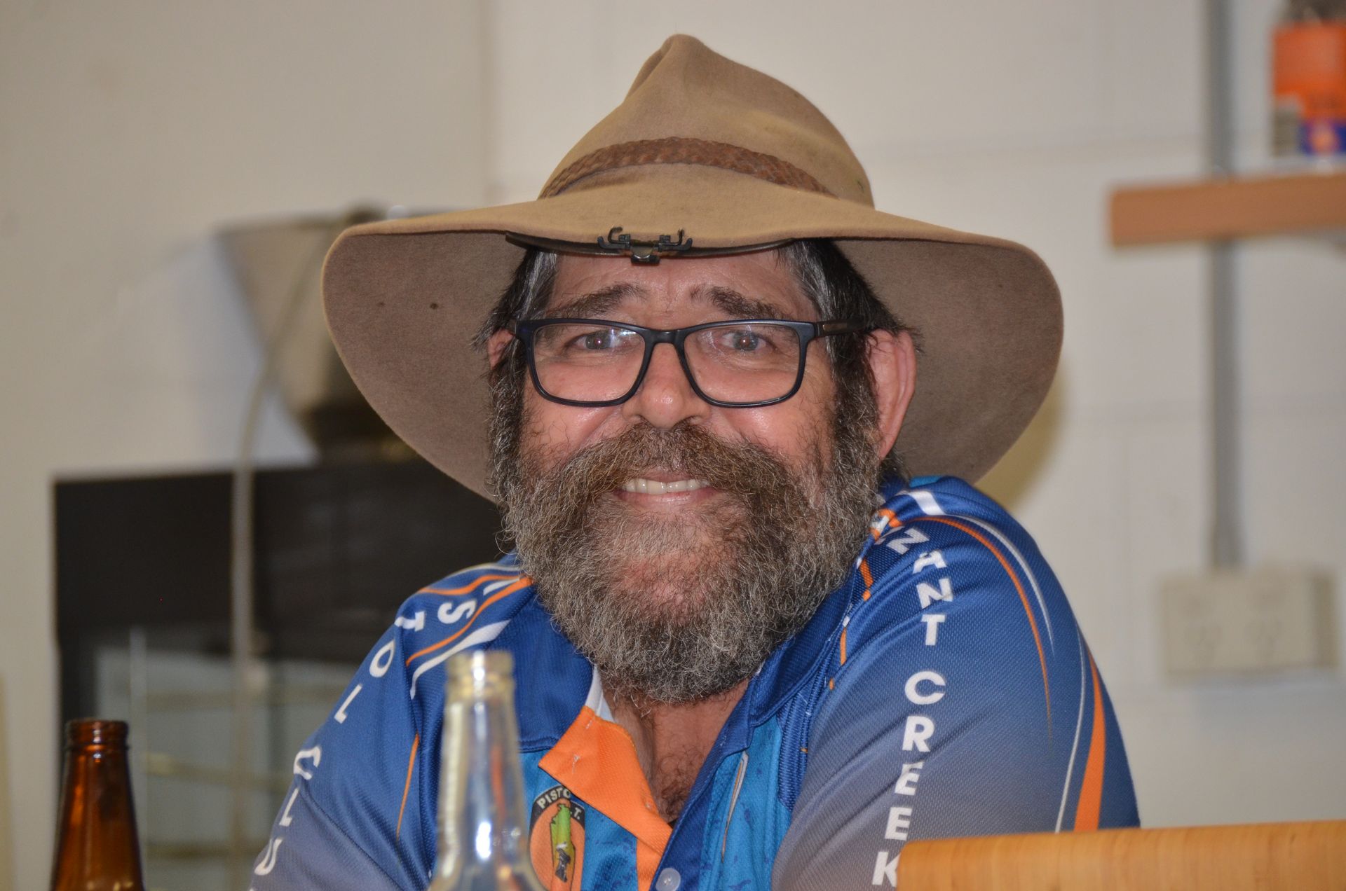 Man wearing a hat and glasses smiles, with a beard and a blue shirt. Indoor setting.