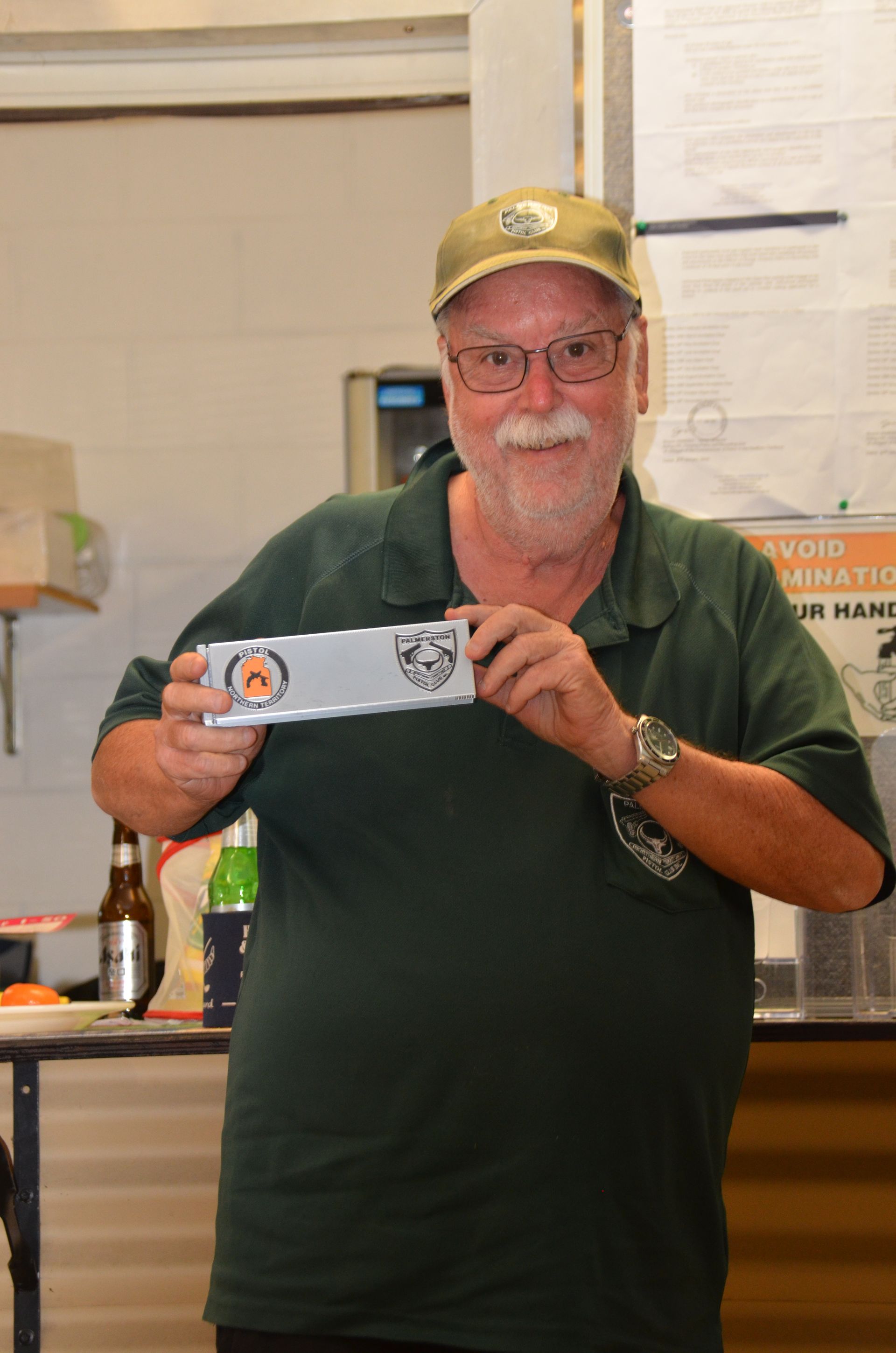 Man with glasses and a cap holds a silver plaque with logos, smiling. Green shirt, indoor setting.
