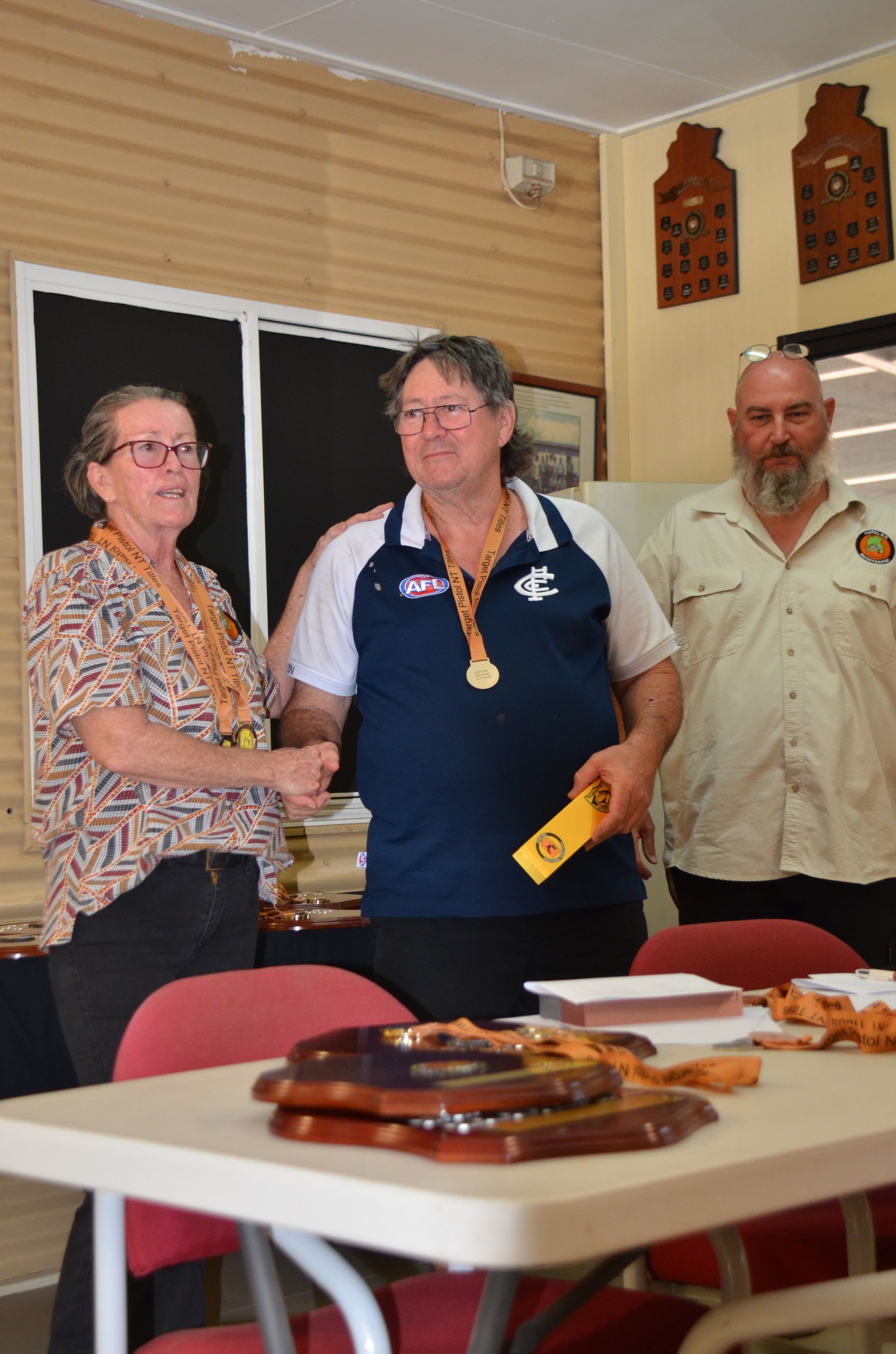 Woman presents a medal to a man in a blue shirt, others watch, plaques on table.