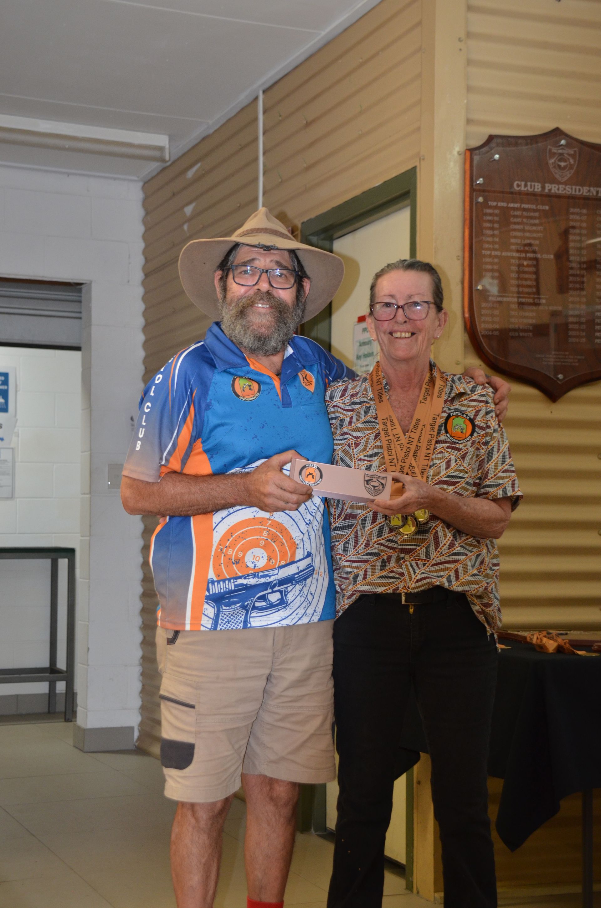 Man in hat and woman smiling, holding an award indoors. Man wears blue shirt, khaki shorts.