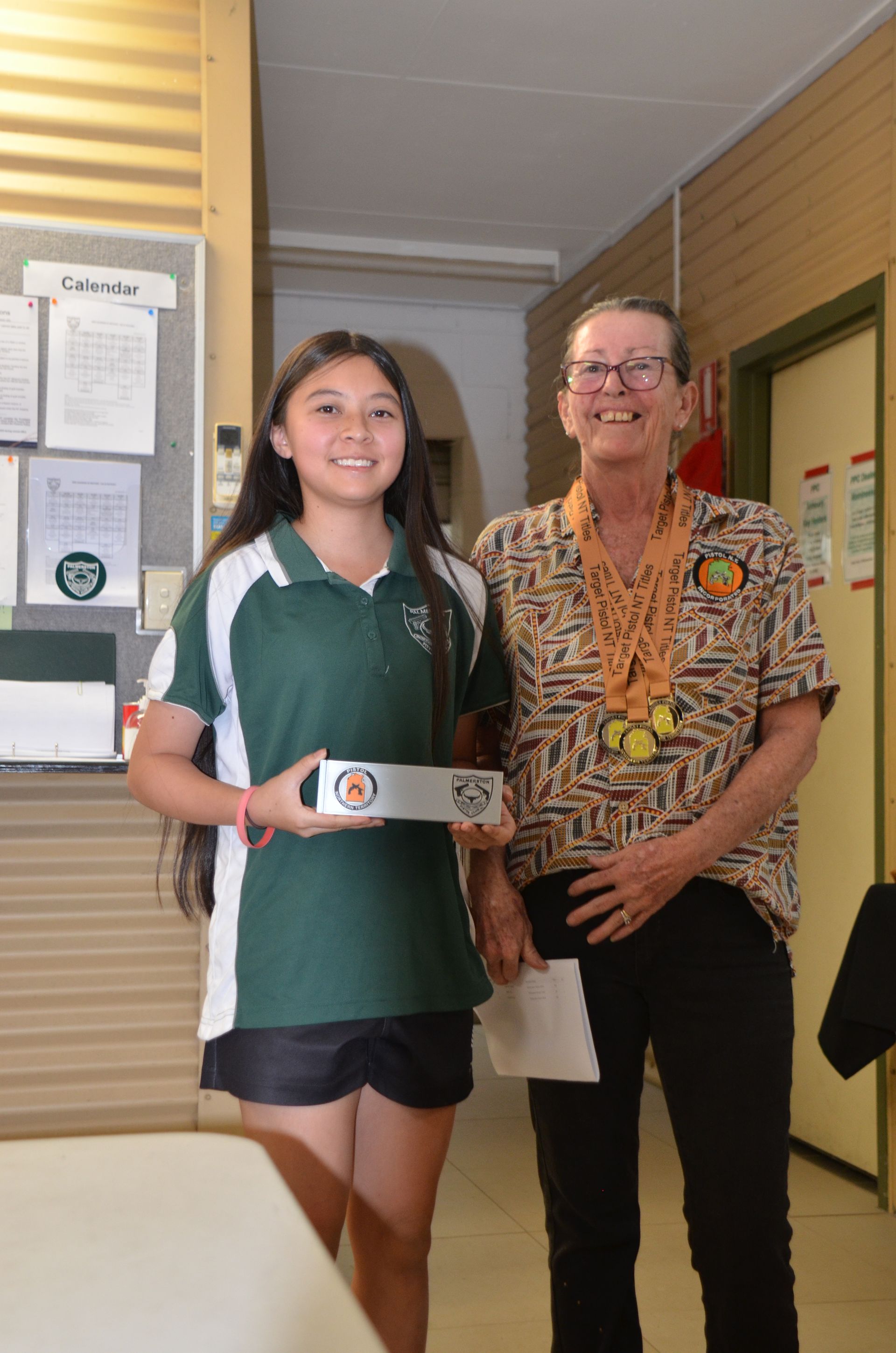 Young woman in school uniform and older woman smiling, posing with an award in a school setting.