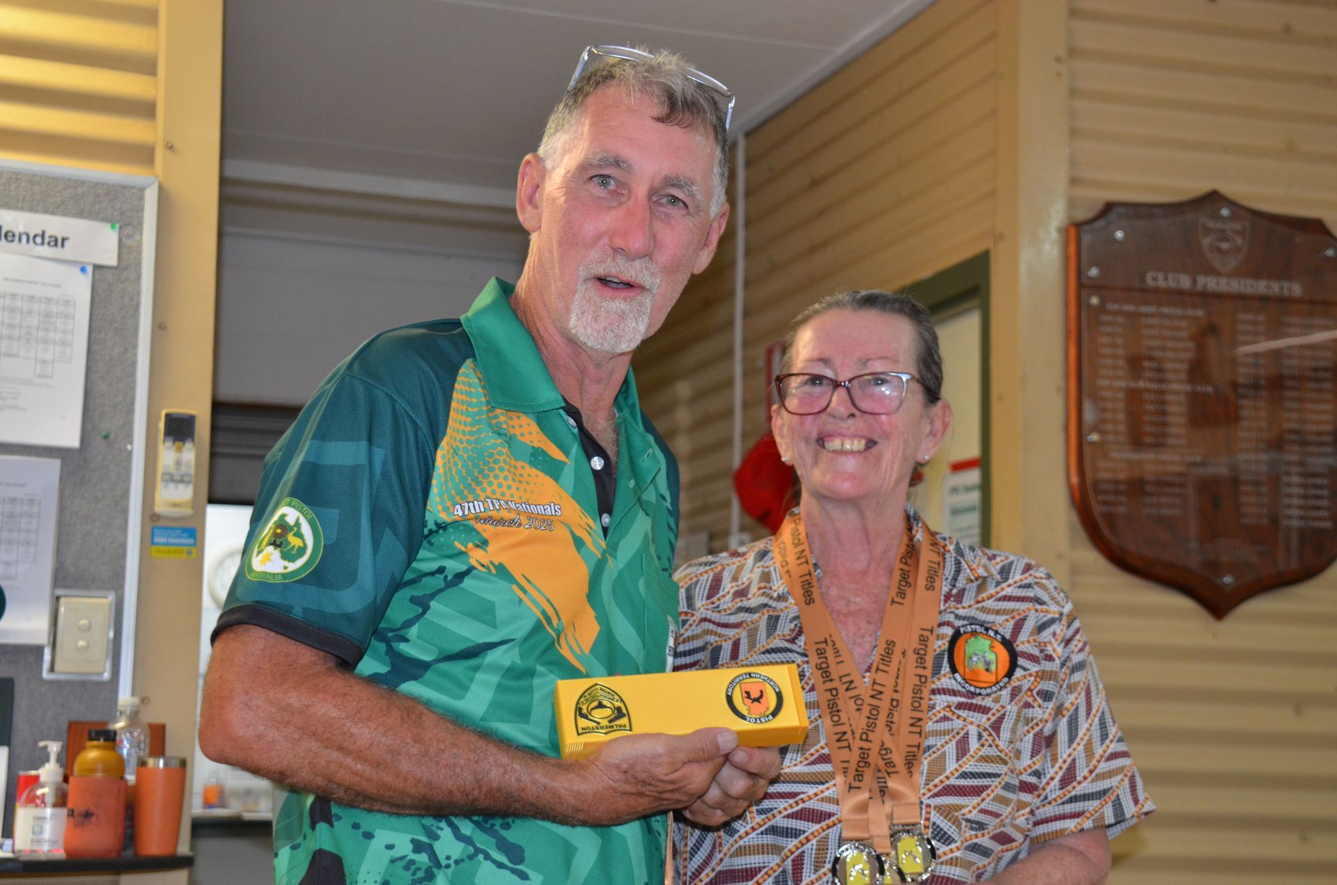 Man in green shirt presents a prize box to smiling woman wearing medals, inside a building.