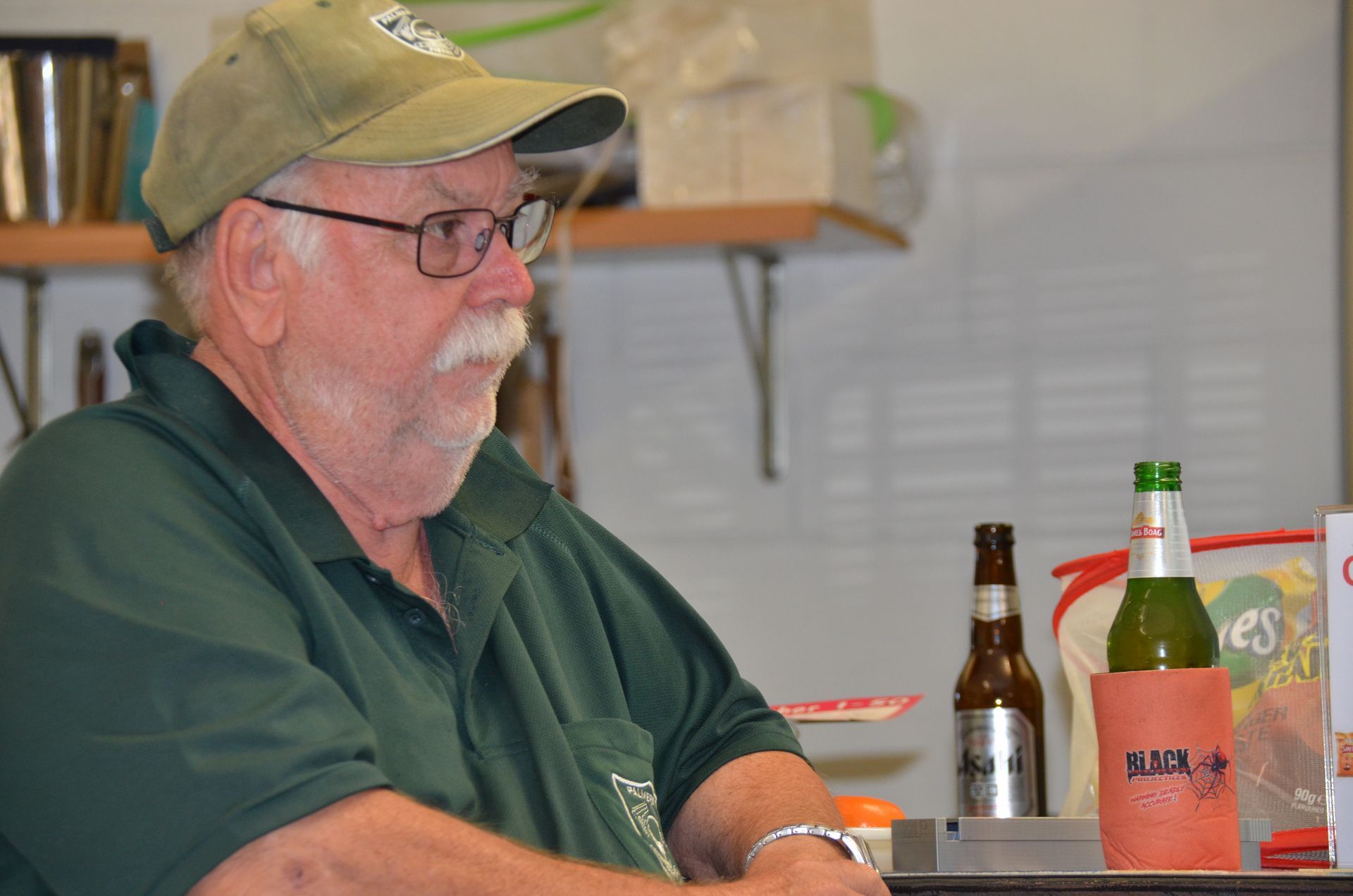 Man with mustache and glasses wearing a green cap and shirt, sitting near two beer bottles.