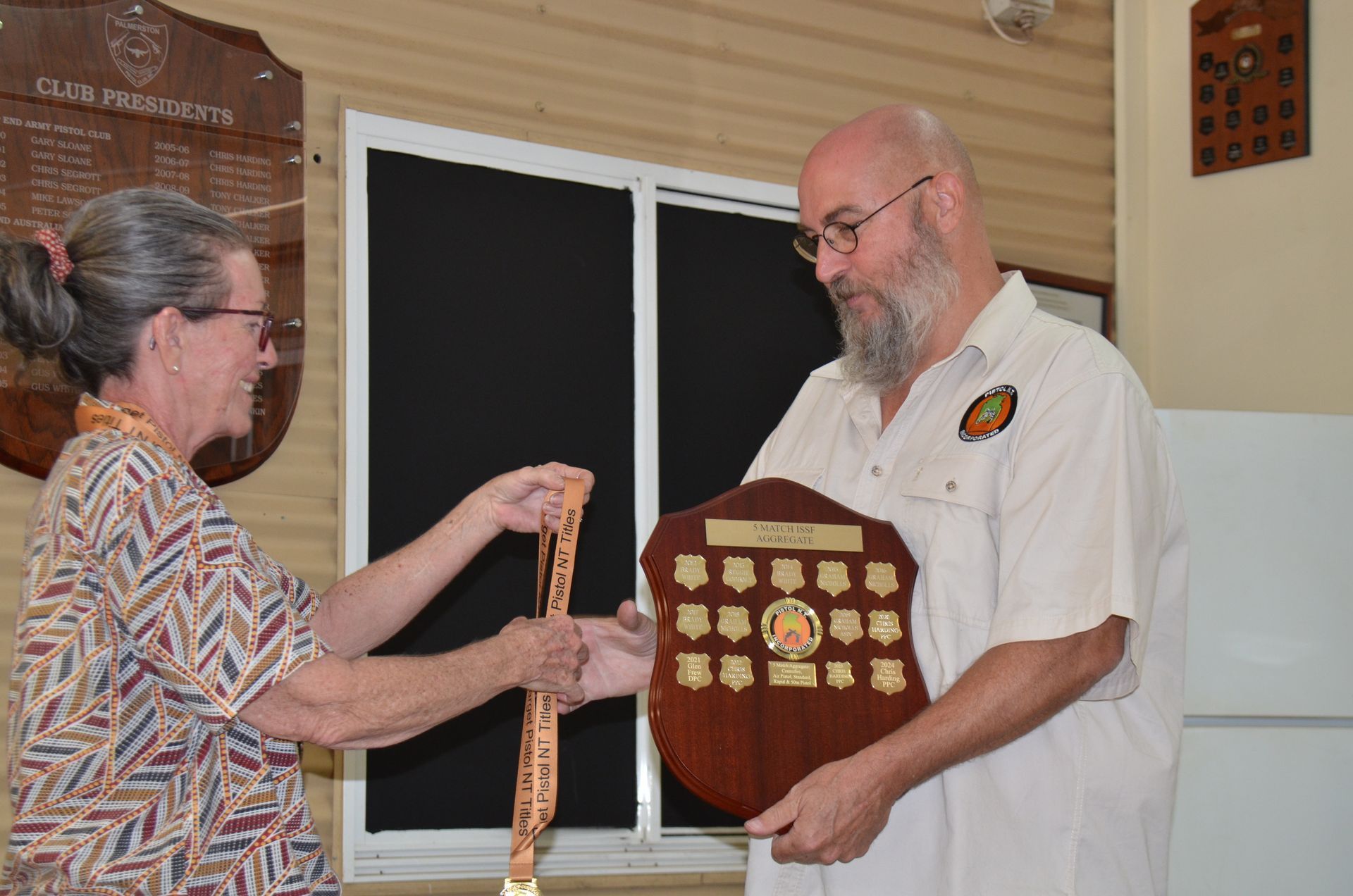 Woman handing a man a medal. Man holds a shield trophy. Indoors.