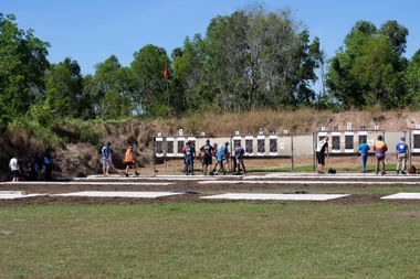 Shooting range with several people in blue and orange shirts aiming at targets, sunny day.