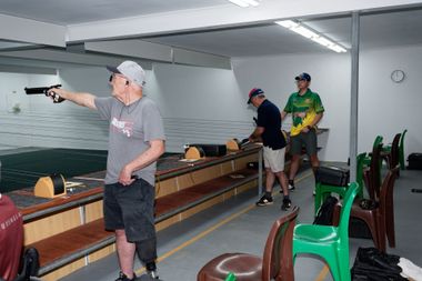 Man aiming pistol at a shooting range, others nearby. Interior, bright lighting, yellow and green chairs.