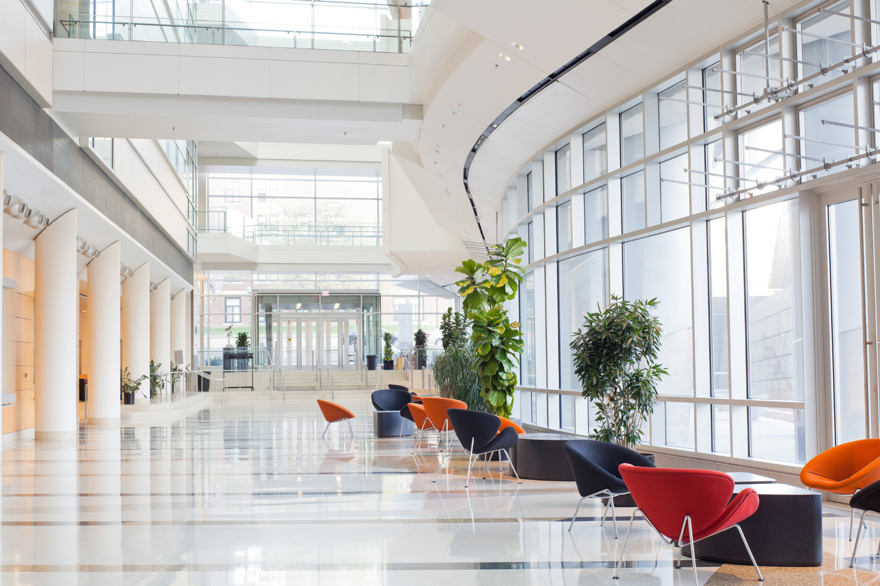 A bright, modern lobby with floor-to-ceiling windows, white pillars, and colorful chairs arranged near indoor plants.