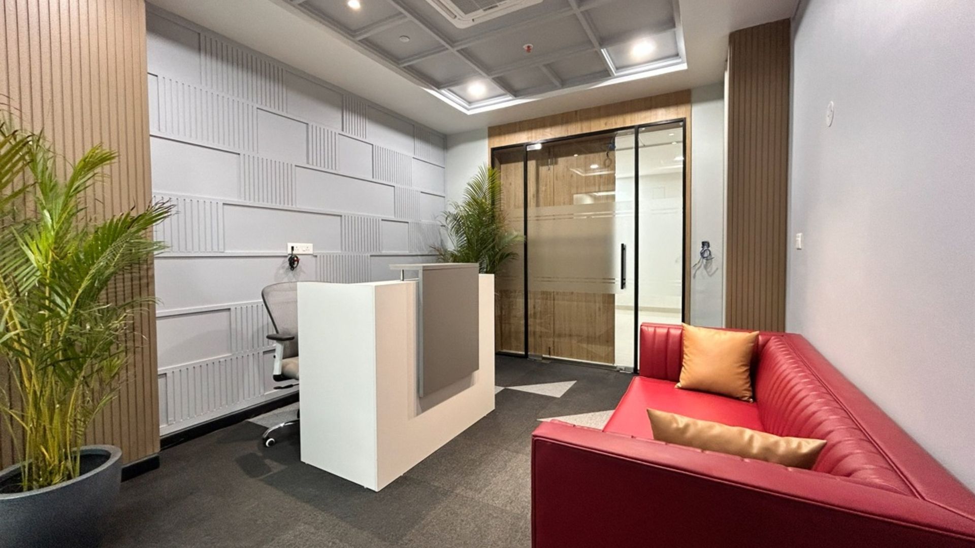 A modern office reception area with a white desk, a red sofa, a potted plant, and a glass-paneled door.