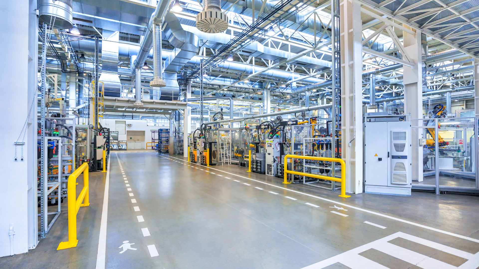 An industrial factory interior with machinery, metallic overhead pipes, and white floor markings for pedestrian pathways.