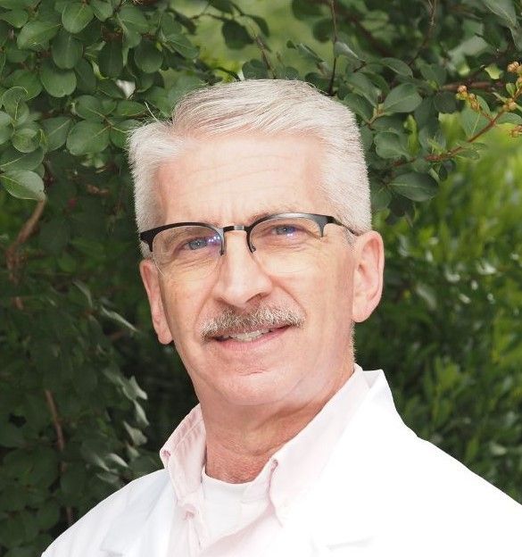 Man with gray hair and mustache wearing glasses and a white coat, smiling.