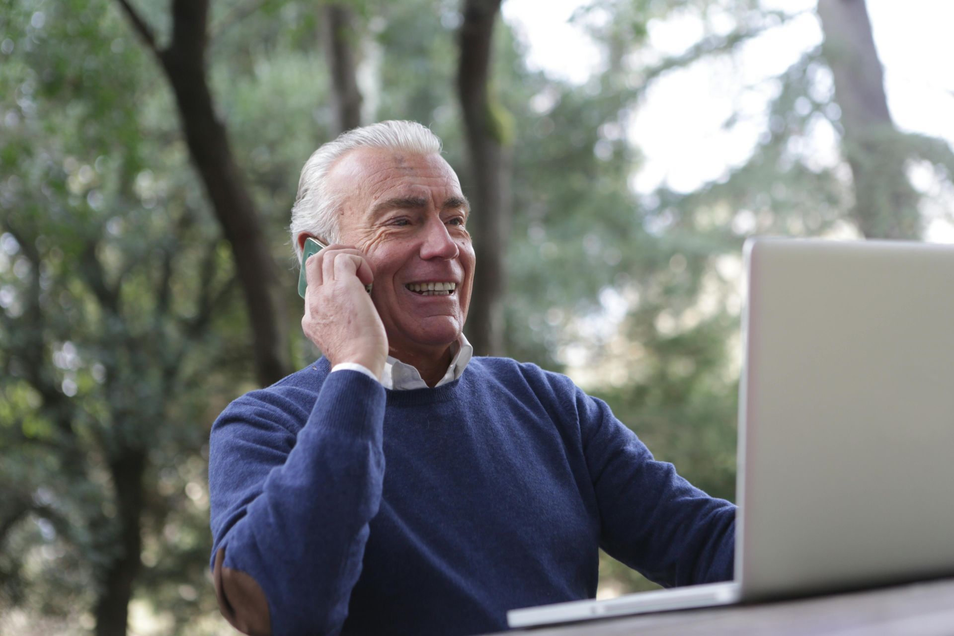 An elderly man is talking on a cell phone while using a laptop computer.