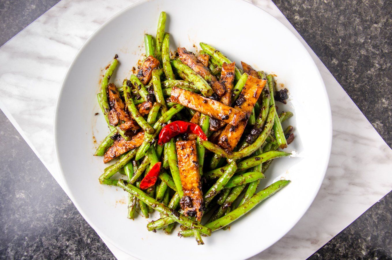 Stir-fried green beans and tofu on a white plate with red chili peppers, seen from above.
