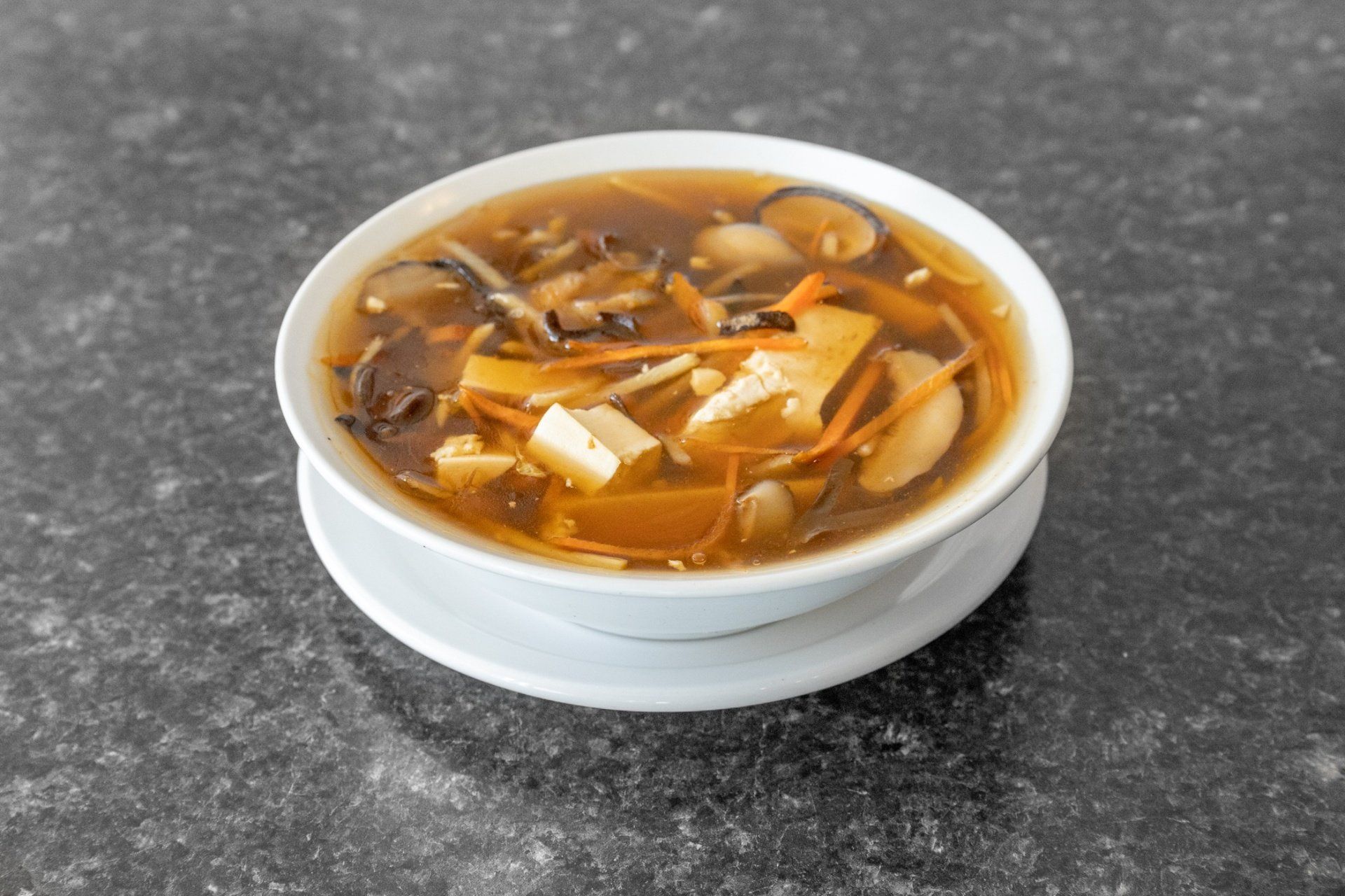 Bowl of hot and sour soup on a white saucer, on a speckled gray surface.
