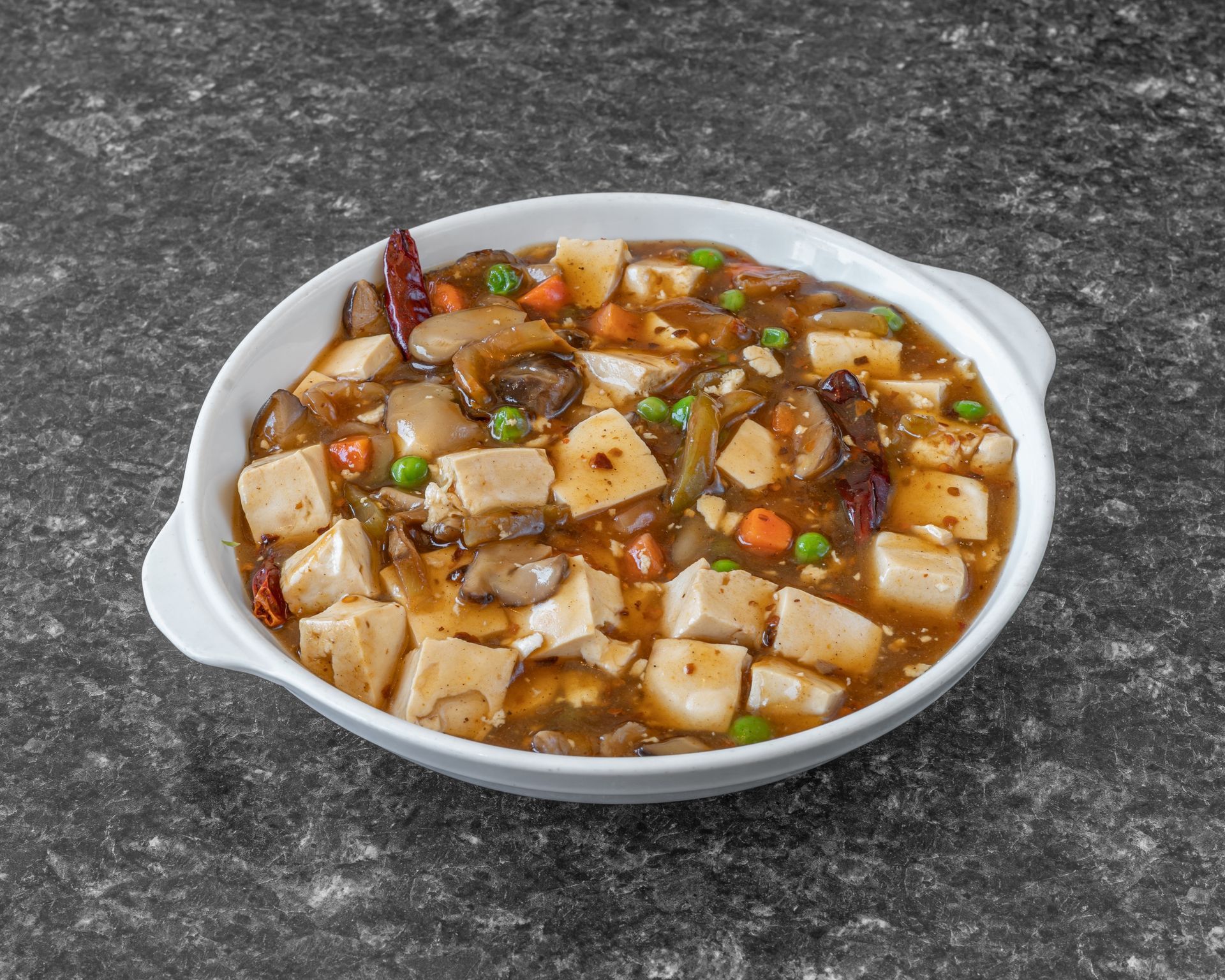 Mapo tofu dish with tofu cubes, mushrooms, peas, and carrots in a brown sauce, white bowl on a gray surface.
