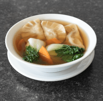 Bowl of dumpling soup with bok choy, carrots, and broth in a white bowl on a gray surface.