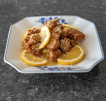 Lemon chicken dish, with sesame seeds, lemon slices, served in a decorative white bowl.