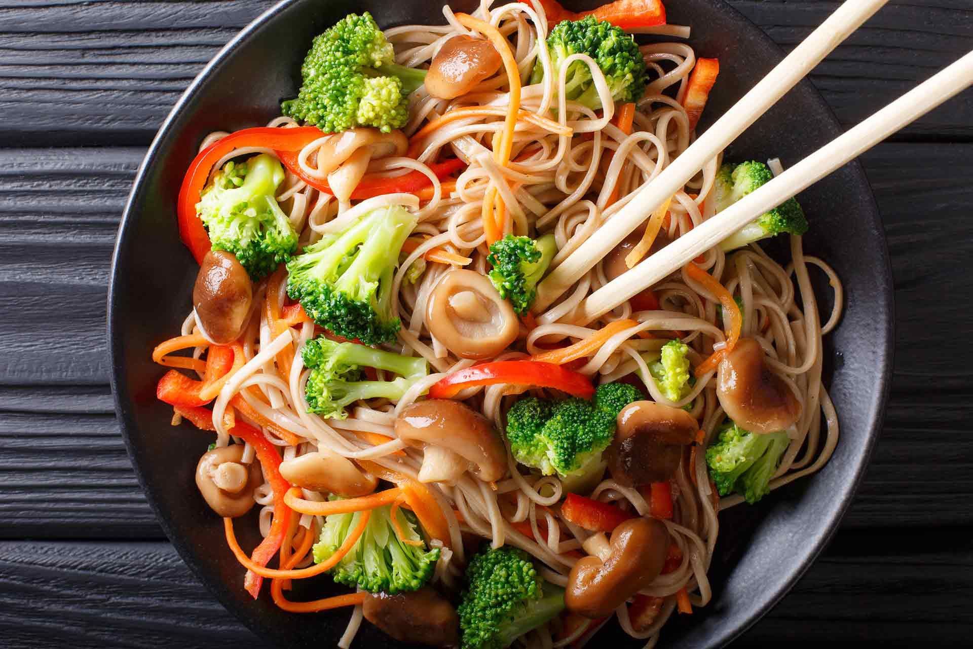 A bowl of stir-fry noodles with vegetables, held with chopsticks.