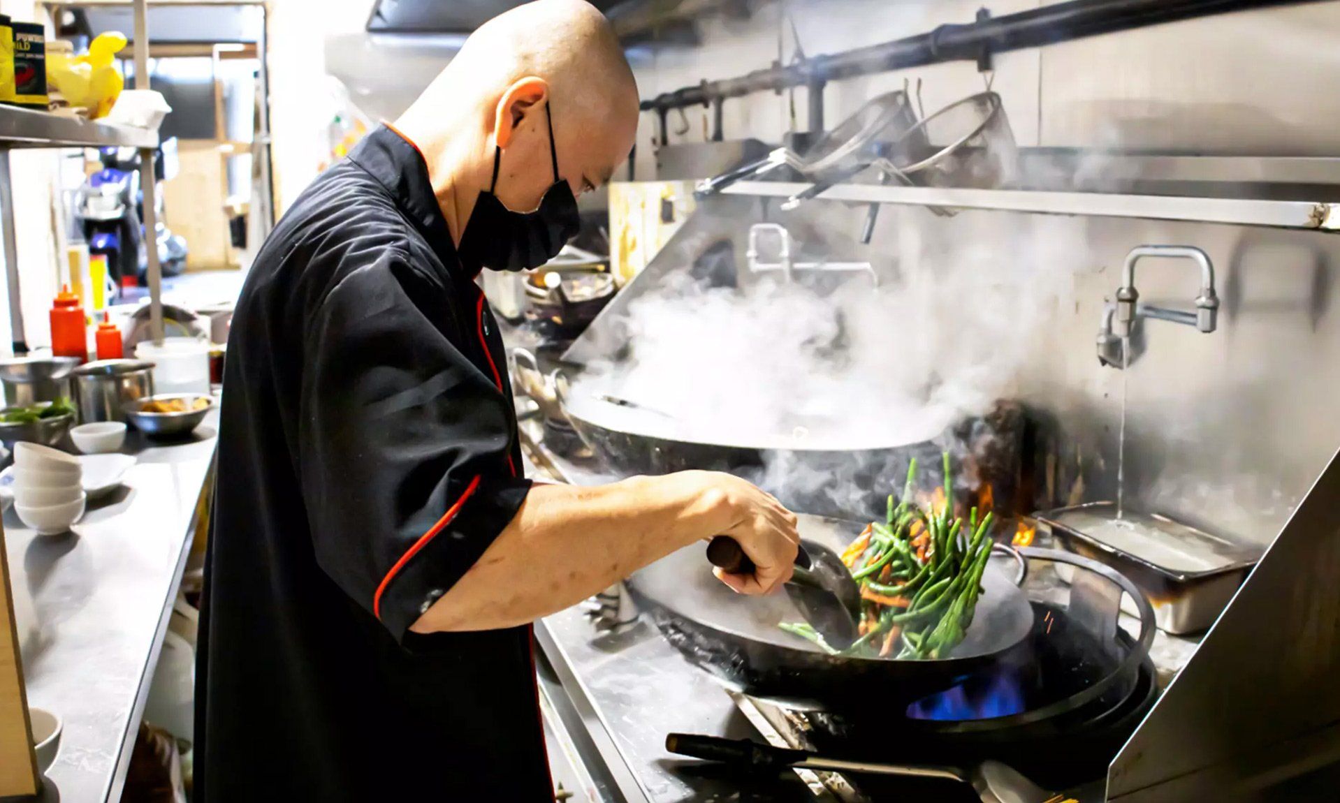 Chef in black uniform and mask stir-fries vegetables in a wok over a blue flame. Kitchen setting.