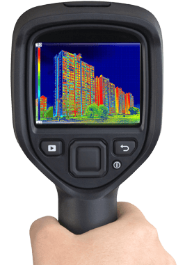 Hand holding a thermal imaging camera pointed at a multi-story building, displaying a color-coded heat map.