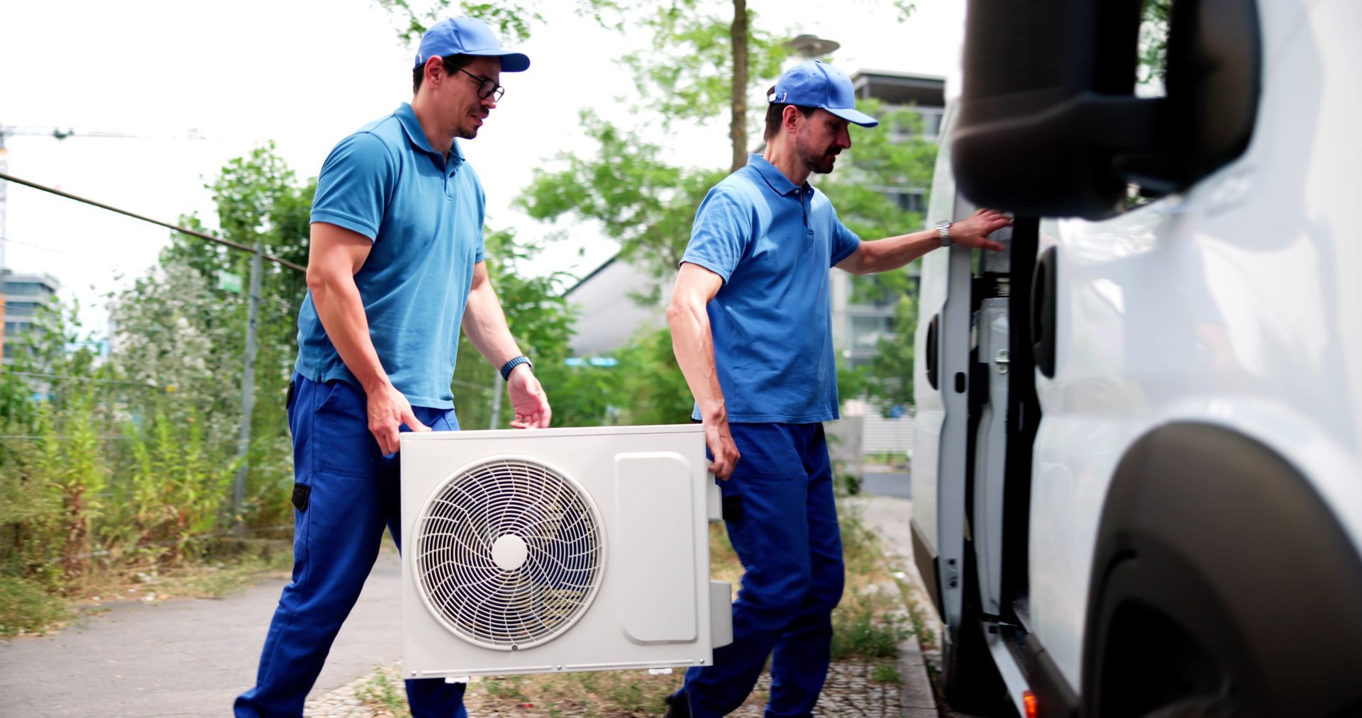 Two workers in blue uniforms load an outdoor unit into a white van roadside. Two workers in blue uniforms load an outdoor unit into a white van roadside.
