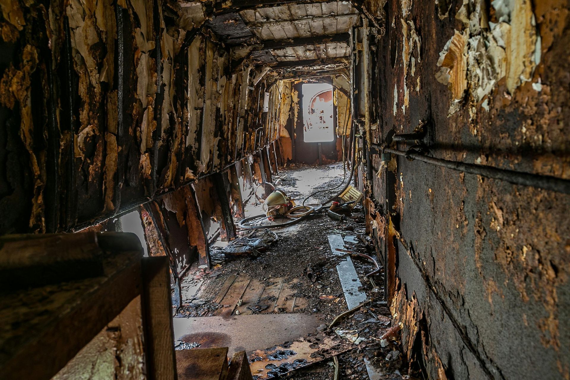 Interior view of a severely damaged, decaying hallway. Debris litters the floor. Walls show signs of fire.
