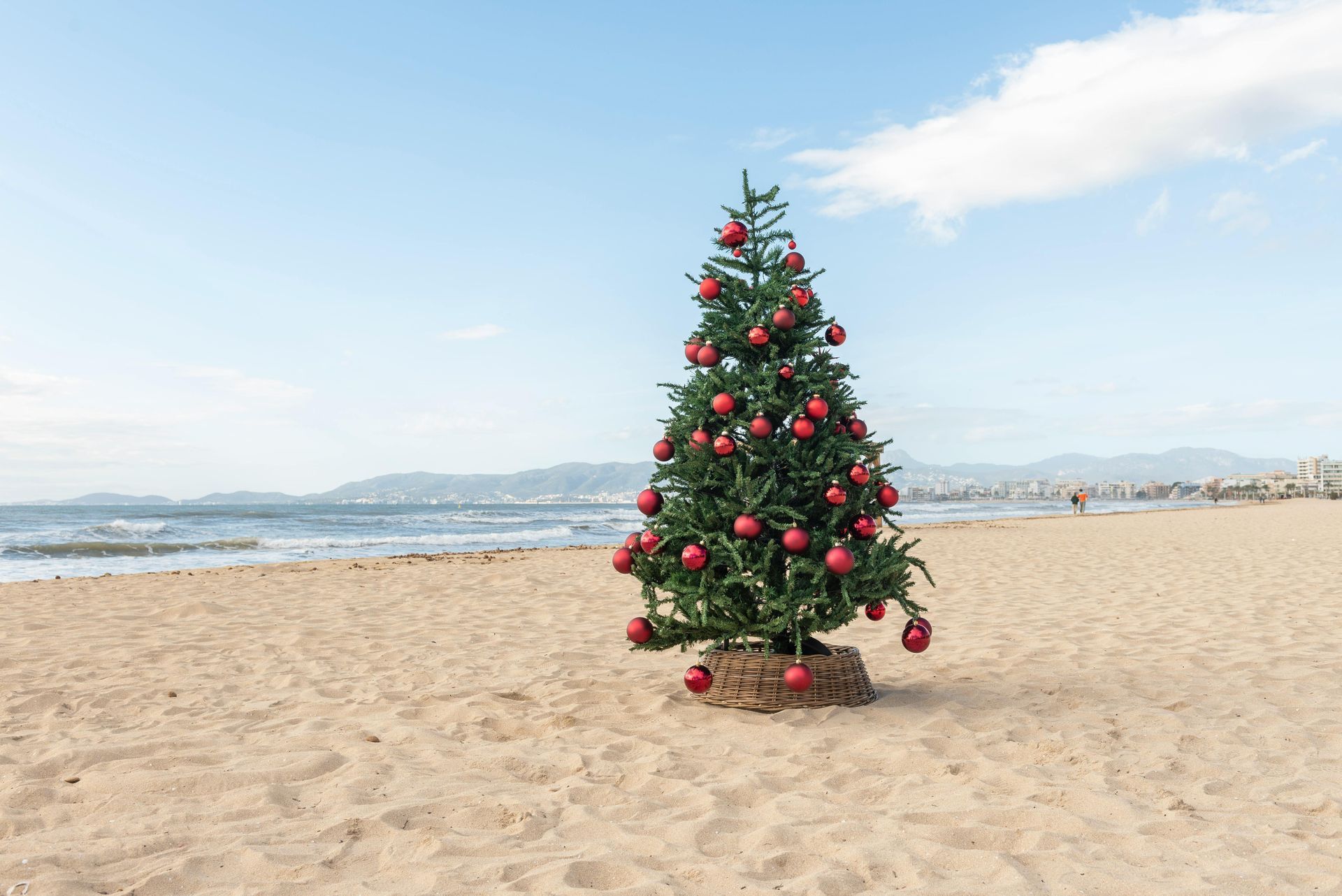 Christmas tree decorated with red ornaments on a sandy beach, with ocean and sky background.