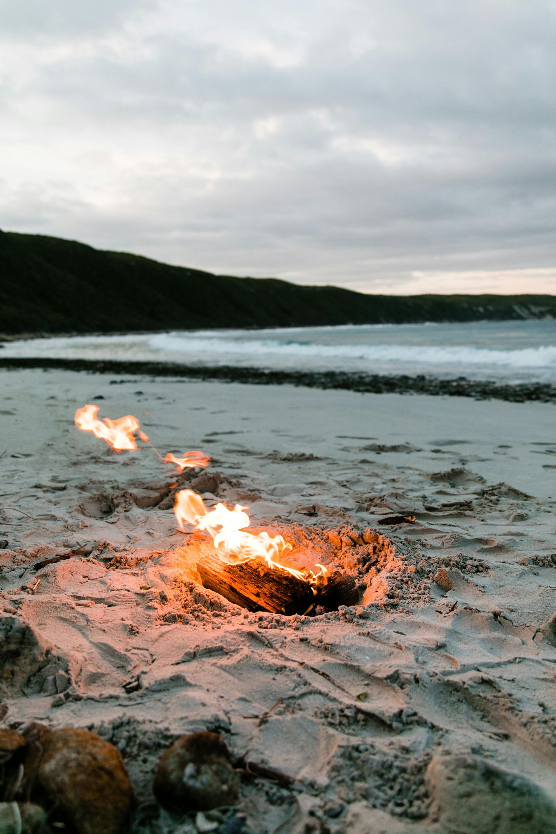 Fire burning on beach sand, waves in the background, cloudy sky.