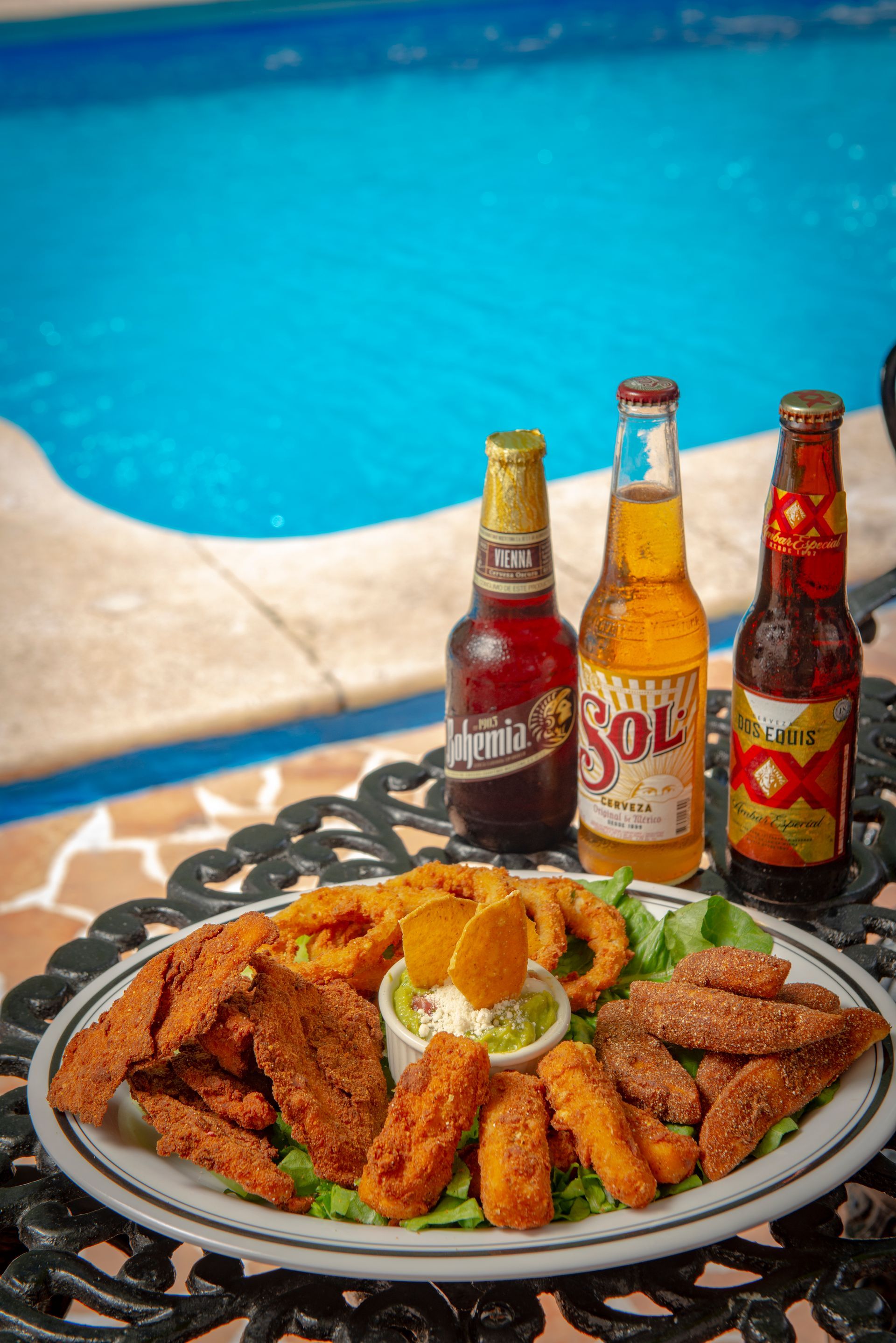 A plate of food and three bottles of beer are on a table next to a pool.