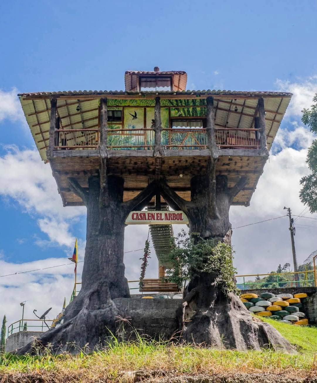 Casa del árbol con balcón de madera, letrero y columpio. Cielo azul de fondo.