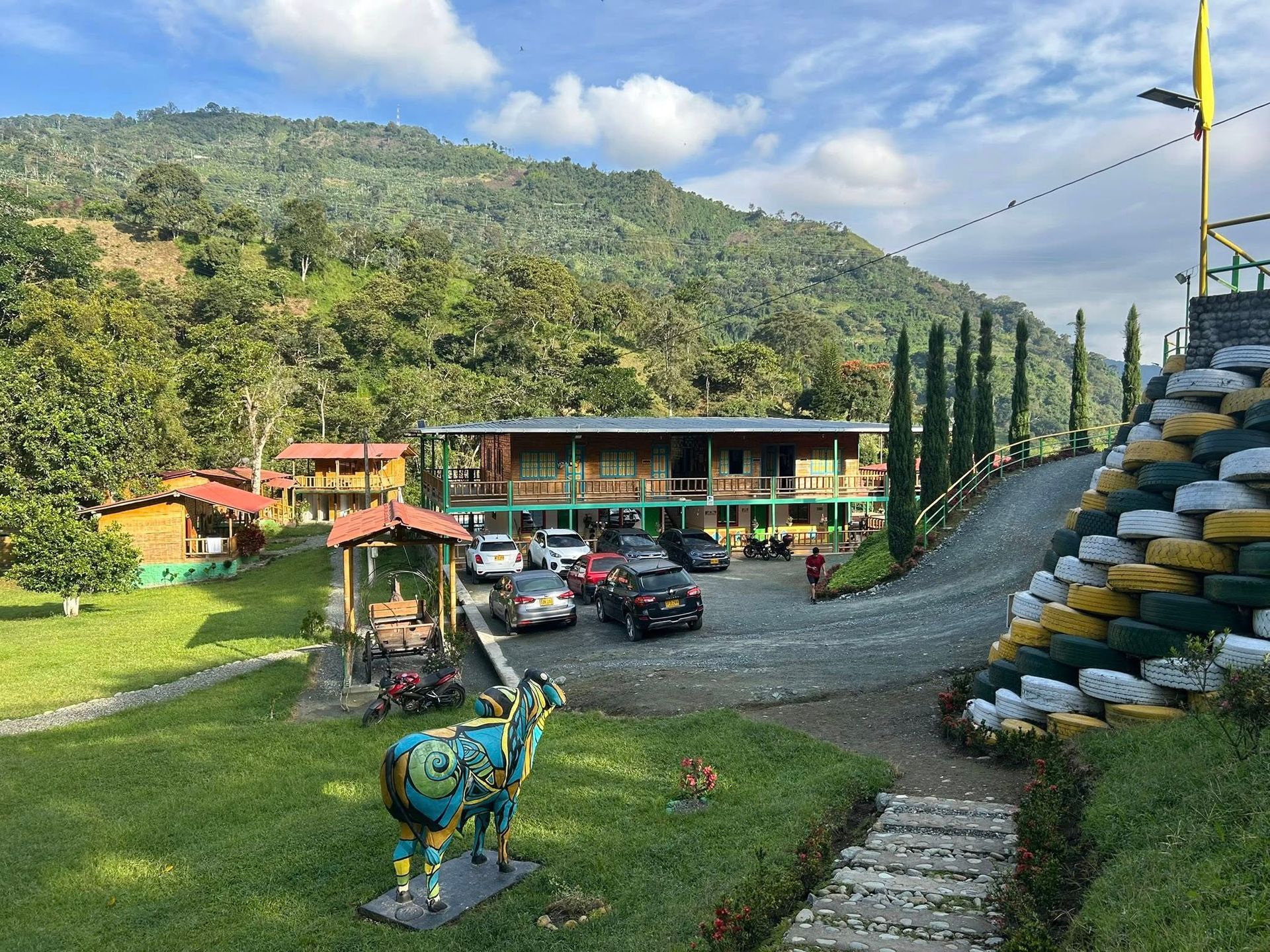 Estación de montaña con edificios, coches, esculturas y árboles bajo un cielo nublado.