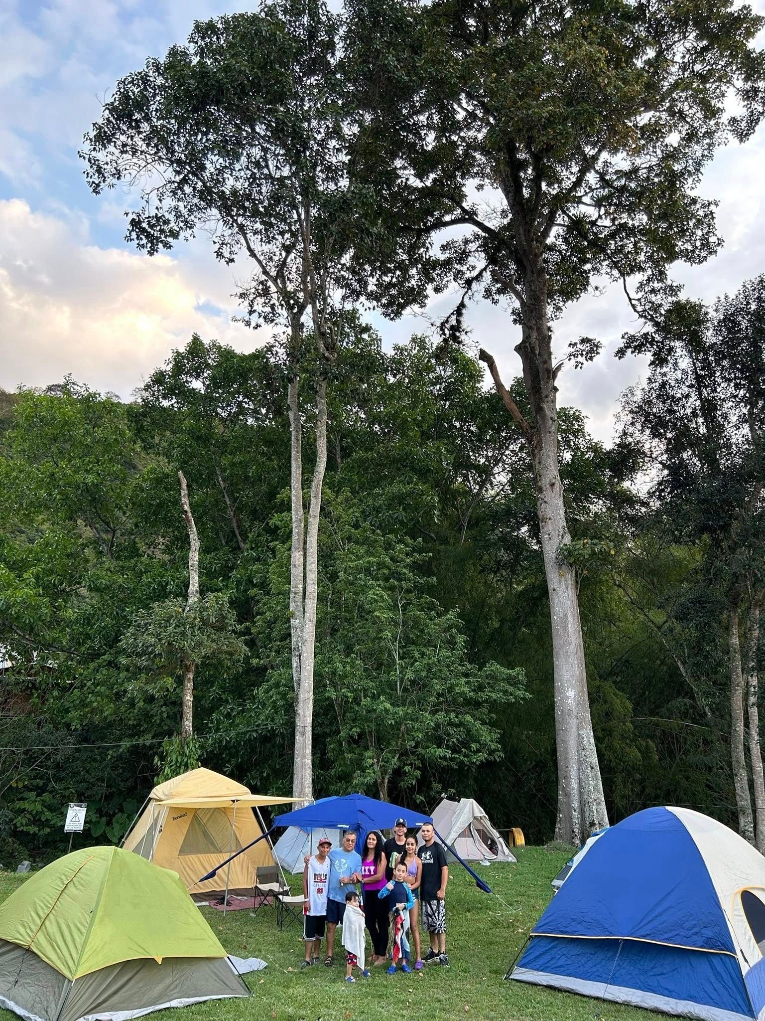 Grupo de personas frente a tiendas de campaña. Exuberantes árboles verdes al fondo.
