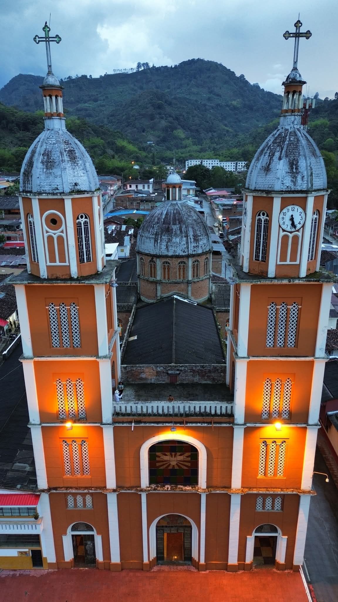 Una iglesia alta de color naranja y blanco con torres gemelas, cruces y una cúpula central con un fondo de montañas.
