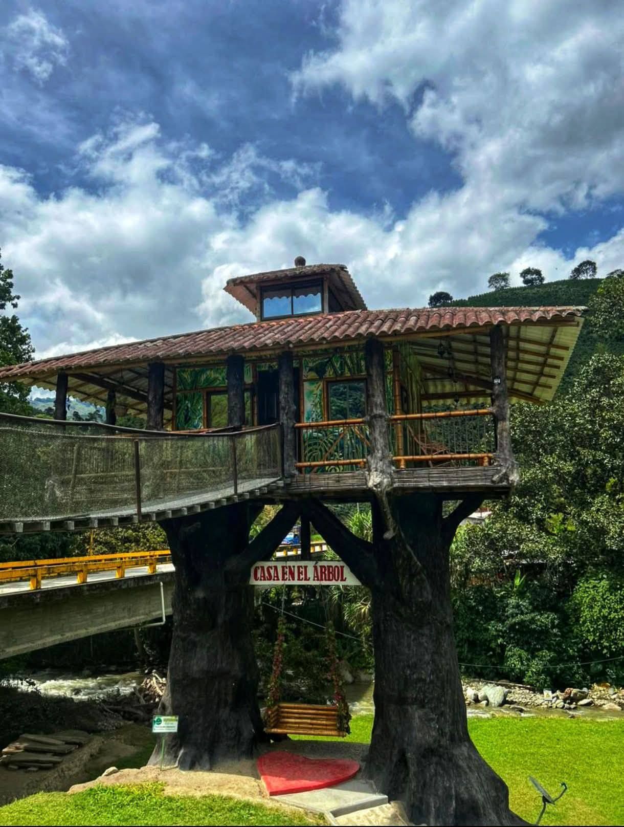 Casa en el árbol con puente, techo de tejas rojas y columpio, bajo un cielo azul nublado.