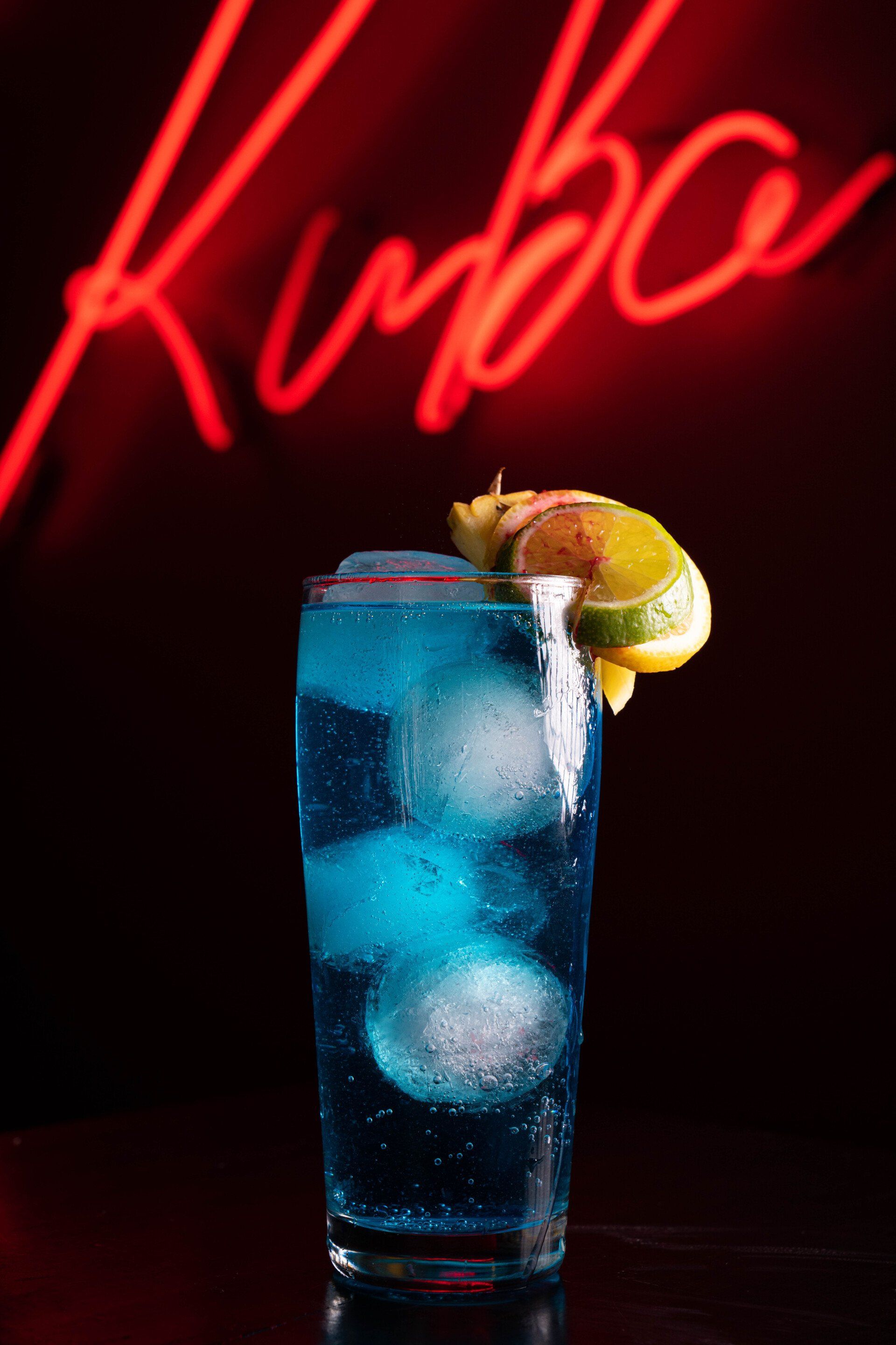 A blue drink with ice and a lemon slice in front of a neon sign that says kuba