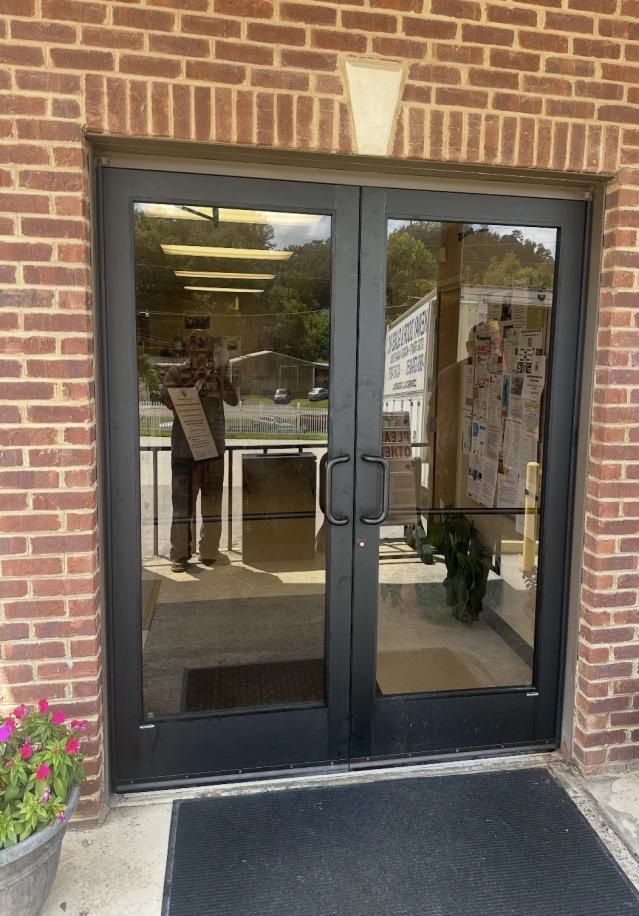 Double glass doors in a brick building. Person reflected in doors holds a sign. Flowers in a pot on the left.