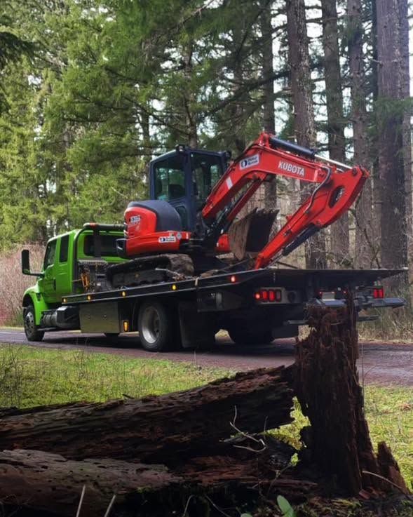 Green flatbed truck hauling a red excavator in a wooded area, with a tree stump in the foreground