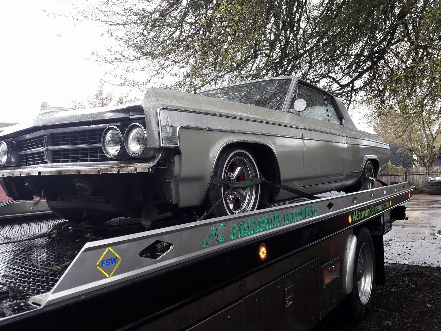 Classic silver car on a tow truck in wet weather under trees