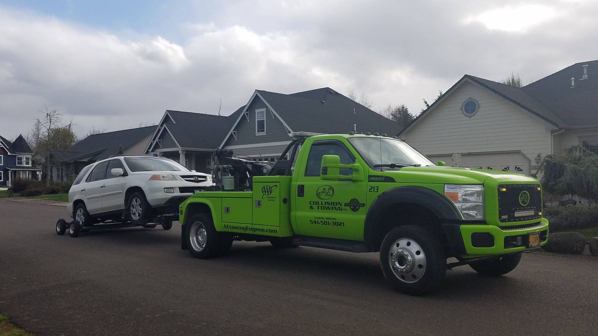Bright green tow truck hauling a white SUV on a residential street under cloudy skies