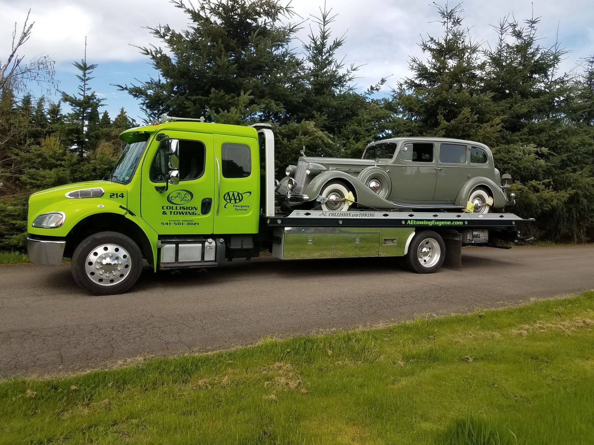 Bright green tow truck carrying a gray vintage car on a flatbed, parked beside a road.