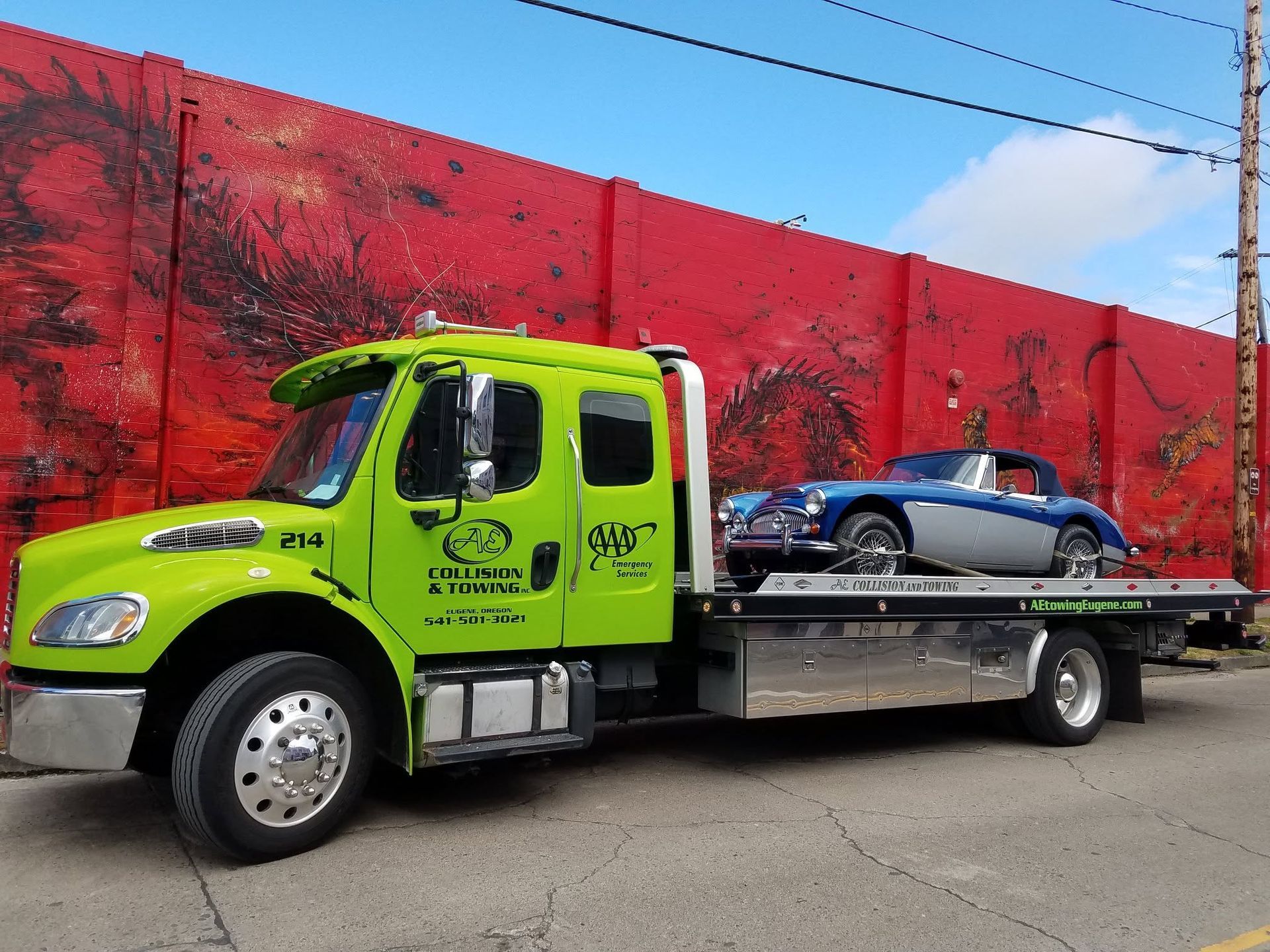 Bright green tow truck hauling a blue car on a flatbed beside a red wall