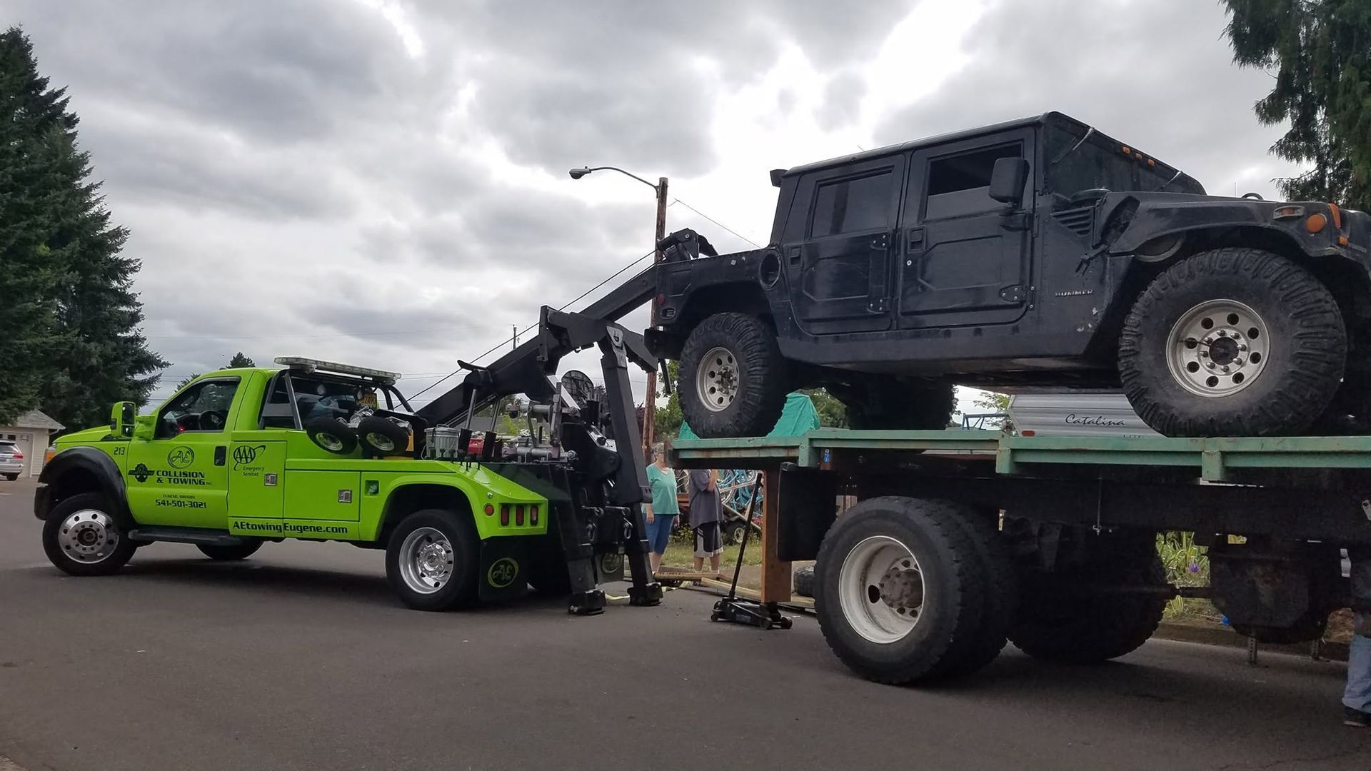 Tow truck loading a black off-road Jeep beside a neon green tow truck under cloudy skies
