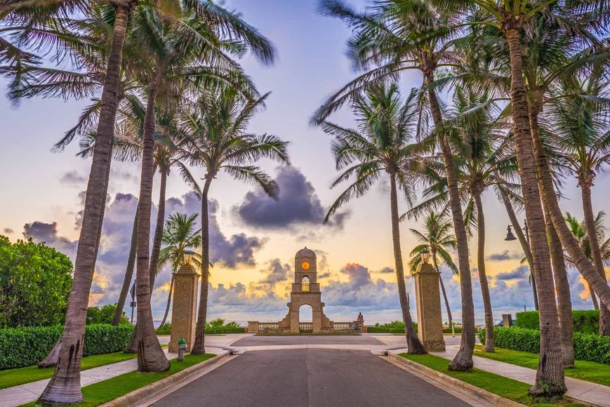 There is a clock tower in the middle of the road surrounded by palm trees.