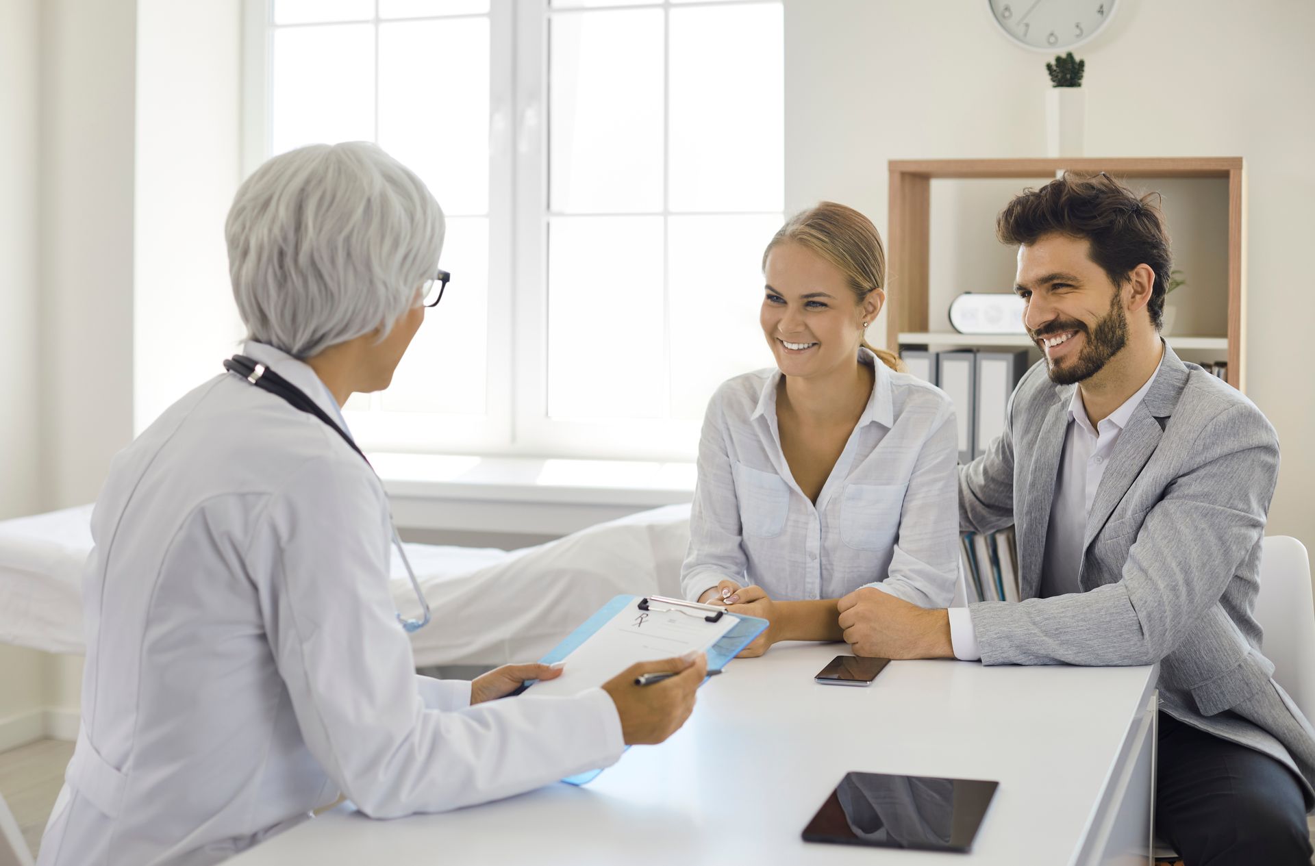 Doctor reviewing documents while consulting with two people seated at a desk in a medical office. Doctor reviewing documents while consulting with two people seated at a desk in a medical office.